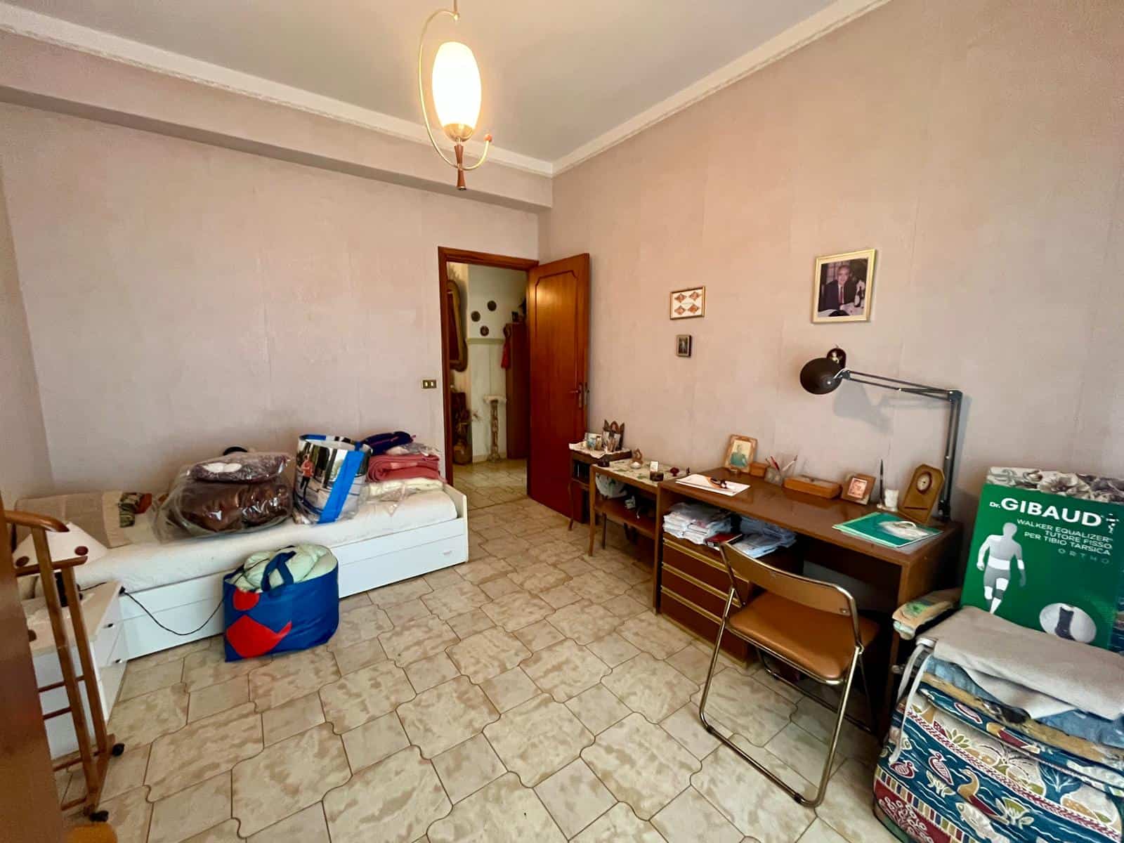 Letto singolo con biancheria e oggetti vari, scrivania con lampada e libri, pareti decorate con quadri, pavimento in piastrelle, ambiente abitabile e funzionale.