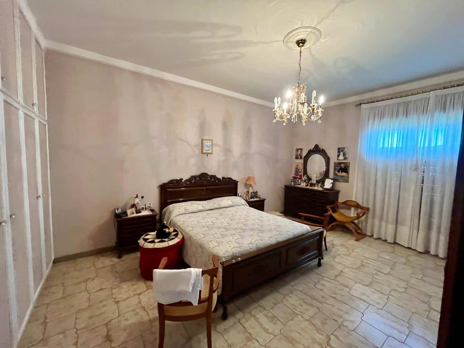 Una stanza da letto spaziosa con letto in legno, lampadario elegante e finestra con tende bianche, arredamento classico e atmosfera accogliente, ideale per case in vendita o affitto.