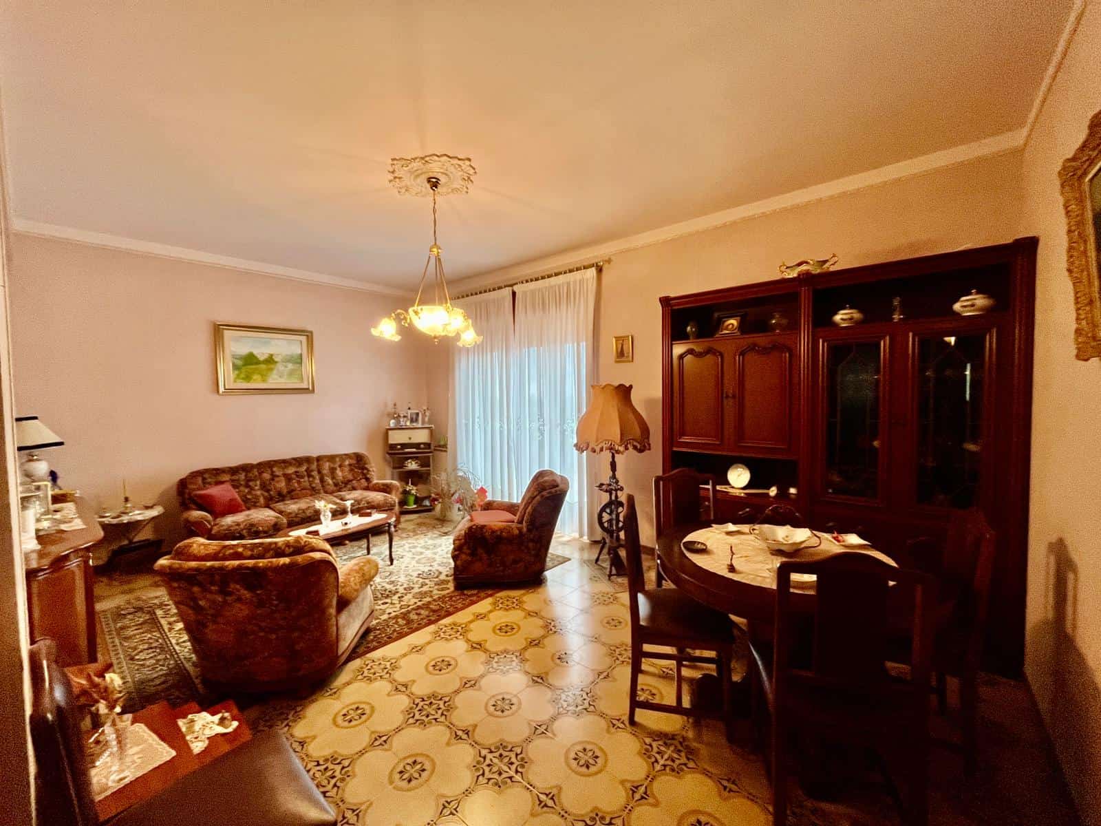 Ampia zona soggiorno con mobili classici, divani e tavolo da pranzo in stile tradizionale, illuminata da lampadario centrale, con pareti decorate e pavimento in piastrelle vintage.