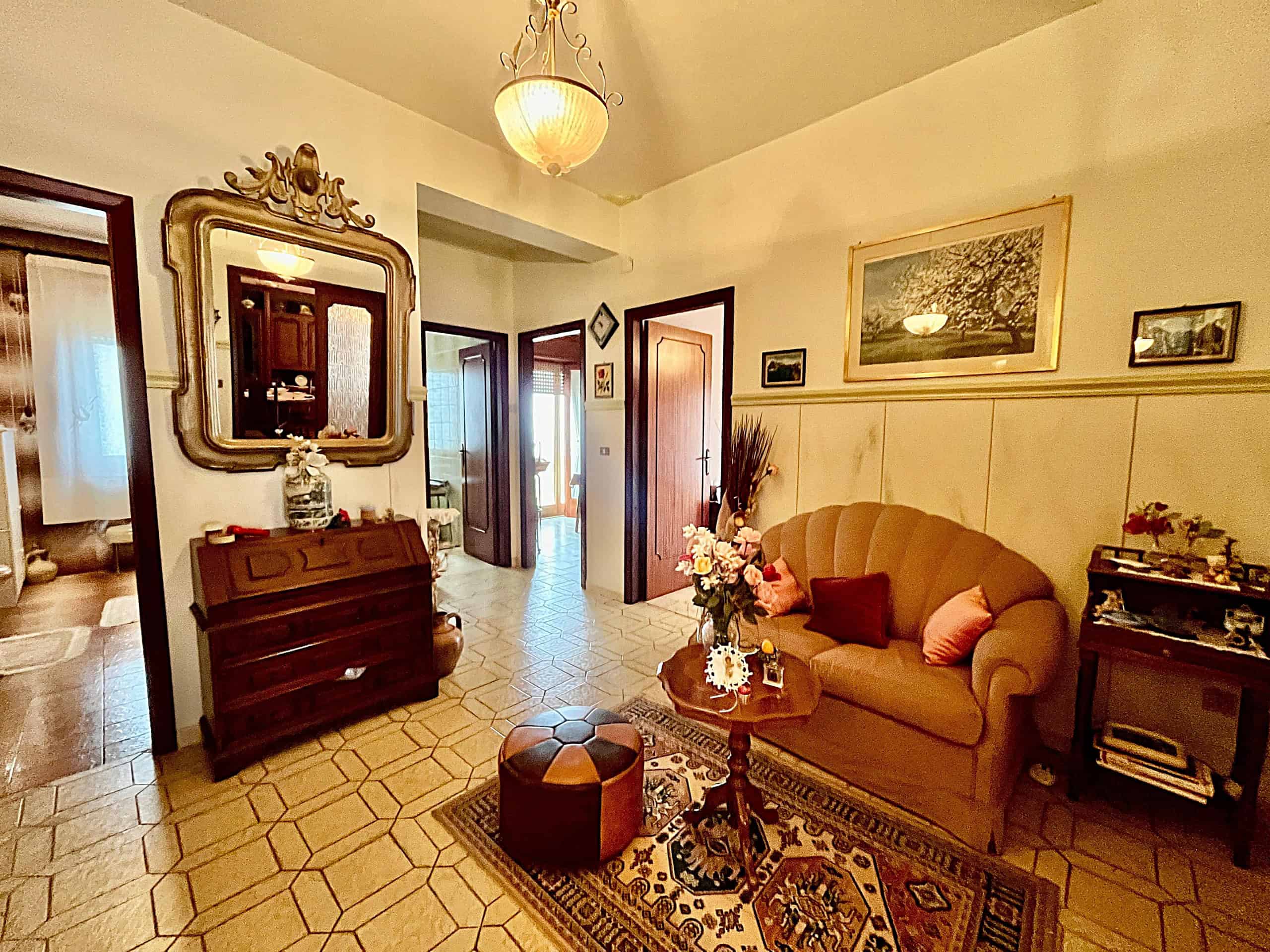 Elegante soggiorno con divano beige, tavolino in legno, e decorazioni floreali, in un ambiente accogliente e ben illuminato, ideale per momenti di relax e convivialità.