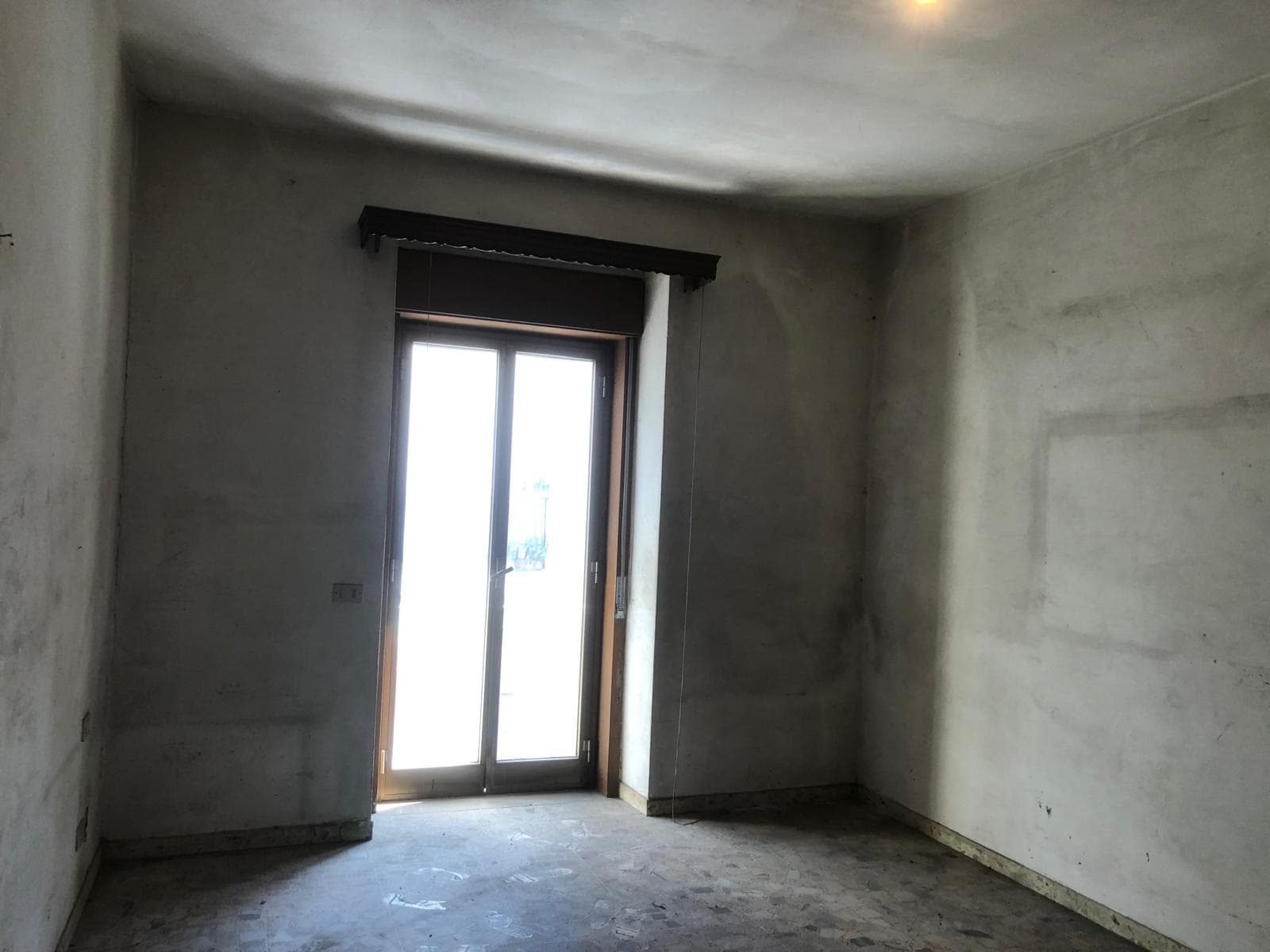 Vetro scorrevole in una stanza vuota in fase di ristrutturazione, con pareti e soffitto in cemento grezzo, luce naturale che entra dalla porta finestra.