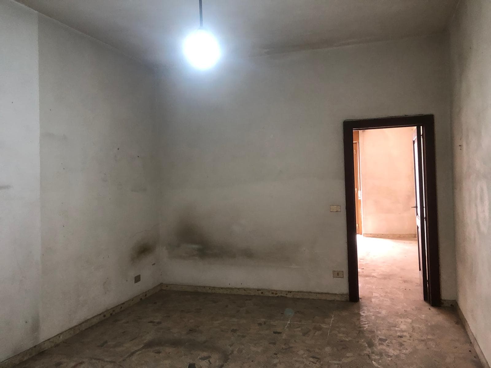 Appartamento vuoto con pareti bianche e pavimento in piastrelle, ingresso in una stanza senza arredamento, illuminata da una lampada a soffitto e luce naturale proveniente da una porta aperta.