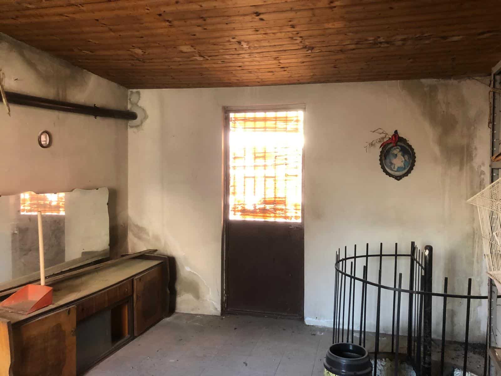 Vecchio locale rustico con pareti bianche, porta in legno e finestra con grate, arredamento semplice e elementi in legno, ideale come spazio di stoccaggio o ristrutturazione.