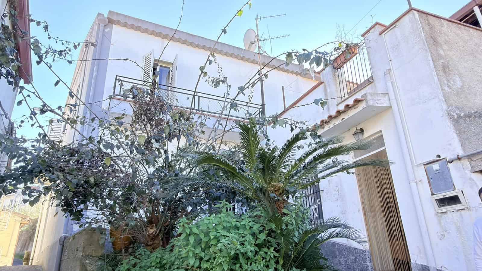 Aggiungi un'immagine di una casa con balcone e piante verdi in un contesto urbano mediterraneo.