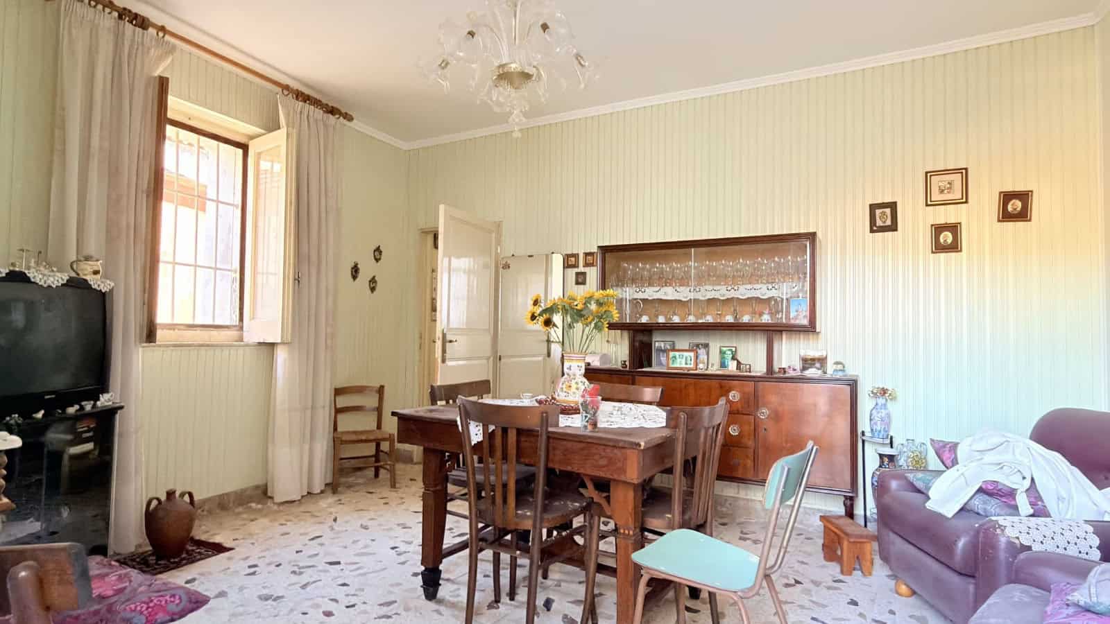 Ampia sala da pranzo in stile classico con tavolo in legno, sedie, credenza e decorazioni vintage, illuminata da luce naturale proveniente dalla finestra, ideale per ambienti familiari e accoglienti.