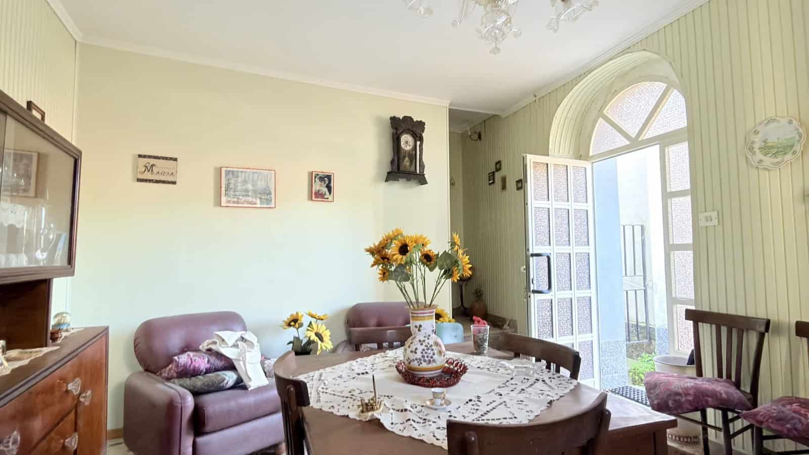 Ampia sala soggiorno con divani in pelle, tavolo da pranzo decorato con fiori freschi, grande finestra ad arco che permette l'ingresso di luce naturale, stile classico e accogliente.