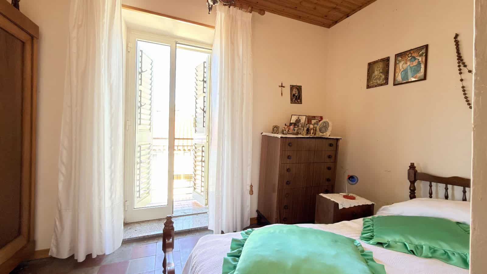 Ampia camera da letto luminosa con balcone, arredi in stile classico, pareti decorate con quadri e oggetti religiosi, ideale per case in vendita o affitto a NextCasa.
