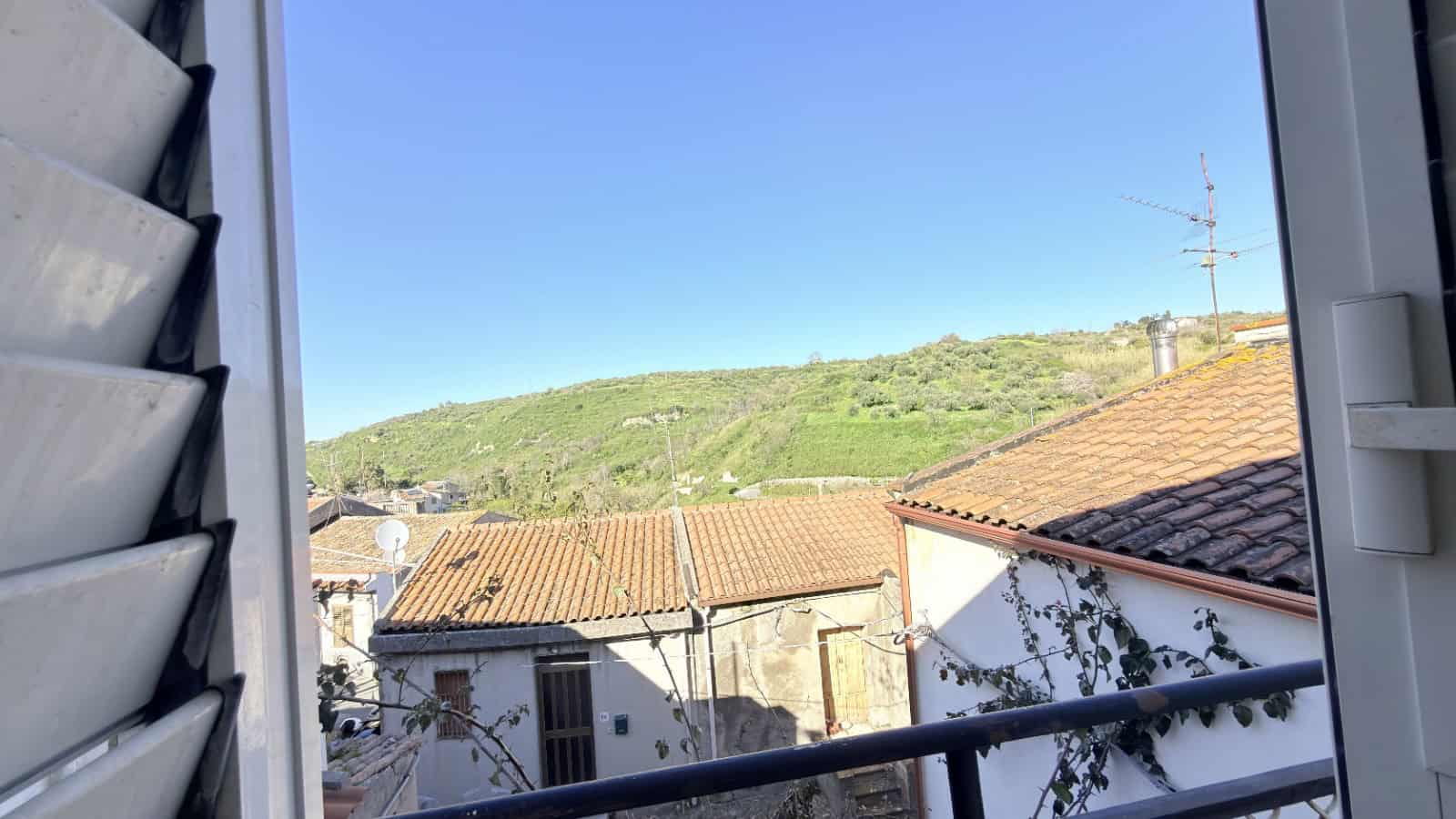 Vista panoramica di un paesaggio collinare con case tradizionali e tetti in terracotta, vista da una finestra con persiane aperte, sotto un cielo sereno e azzurro, ideale per immagini di immobili e pa.