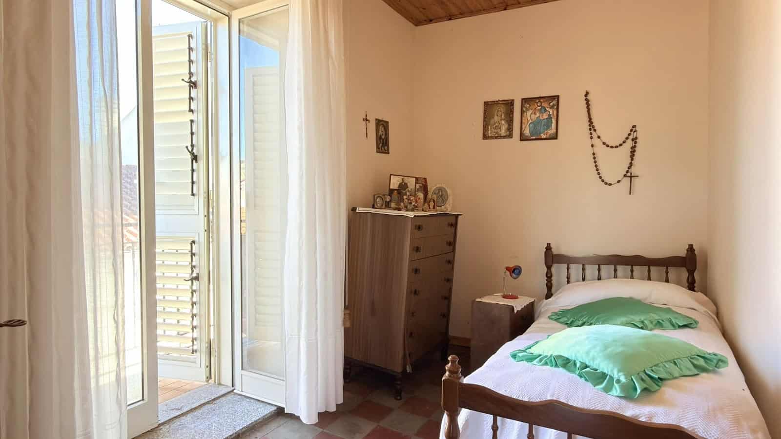 Letto singolo con cuscini verdi in una camera luminosa con pareti beige, finestra con tende leggere e accessori decorativi alle pareti, ambiente accogliente e tranquillo.