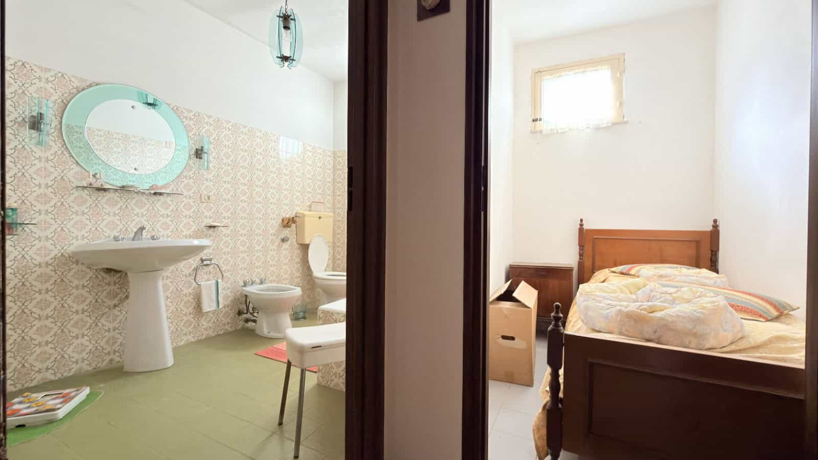 Lavabo e bagno con piastrelle decorative, vista parziale della camera da letto con letto in legno e finestra naturale, ambiente accogliente e ben illuminato.