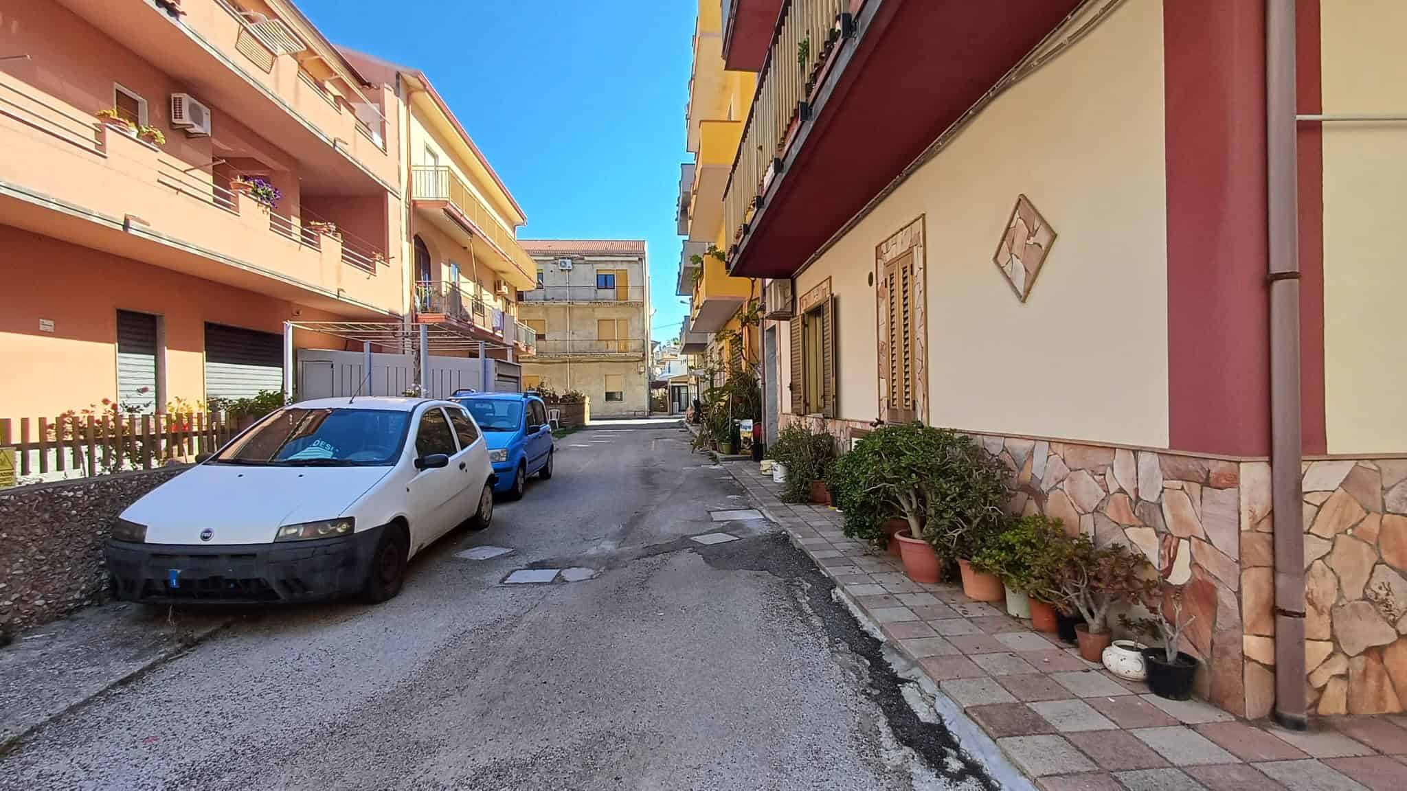 Auto in sosta lungo strada residenziale con edifici multifamiliari, piante in vaso e facciate colorate, in un quartiere urbano soleggiato e tranquillo, ideale per vivere in città.