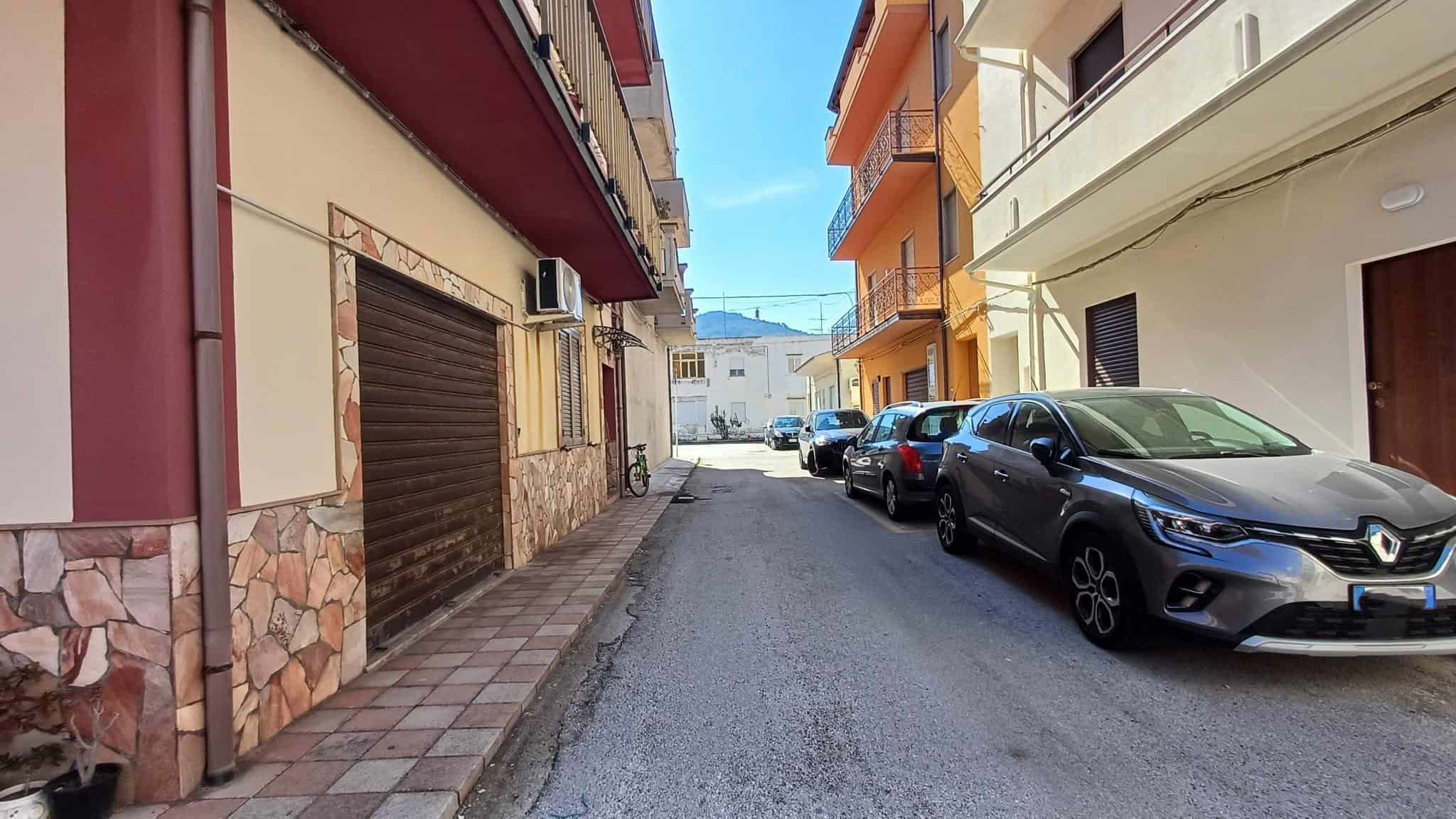 Ampia strada urbana con veicoli parcheggiati, edifici residenziali colorati e cielo sereno, rappresentativa di un quartiere tranquillo e ben servito in Italia.
