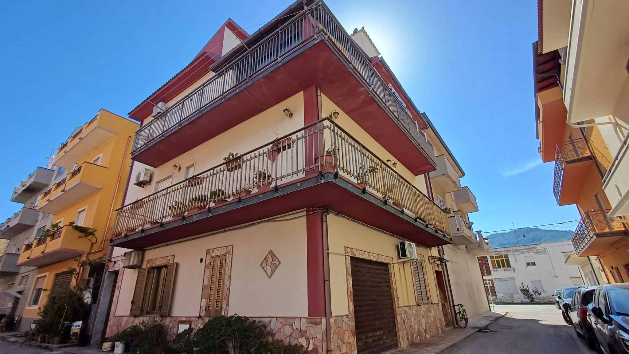 Balcone di un edificio residenziale in zona urbana, con ringhiera in metallo e piante decorative, sotto un cielo azzurro con sole brillante.