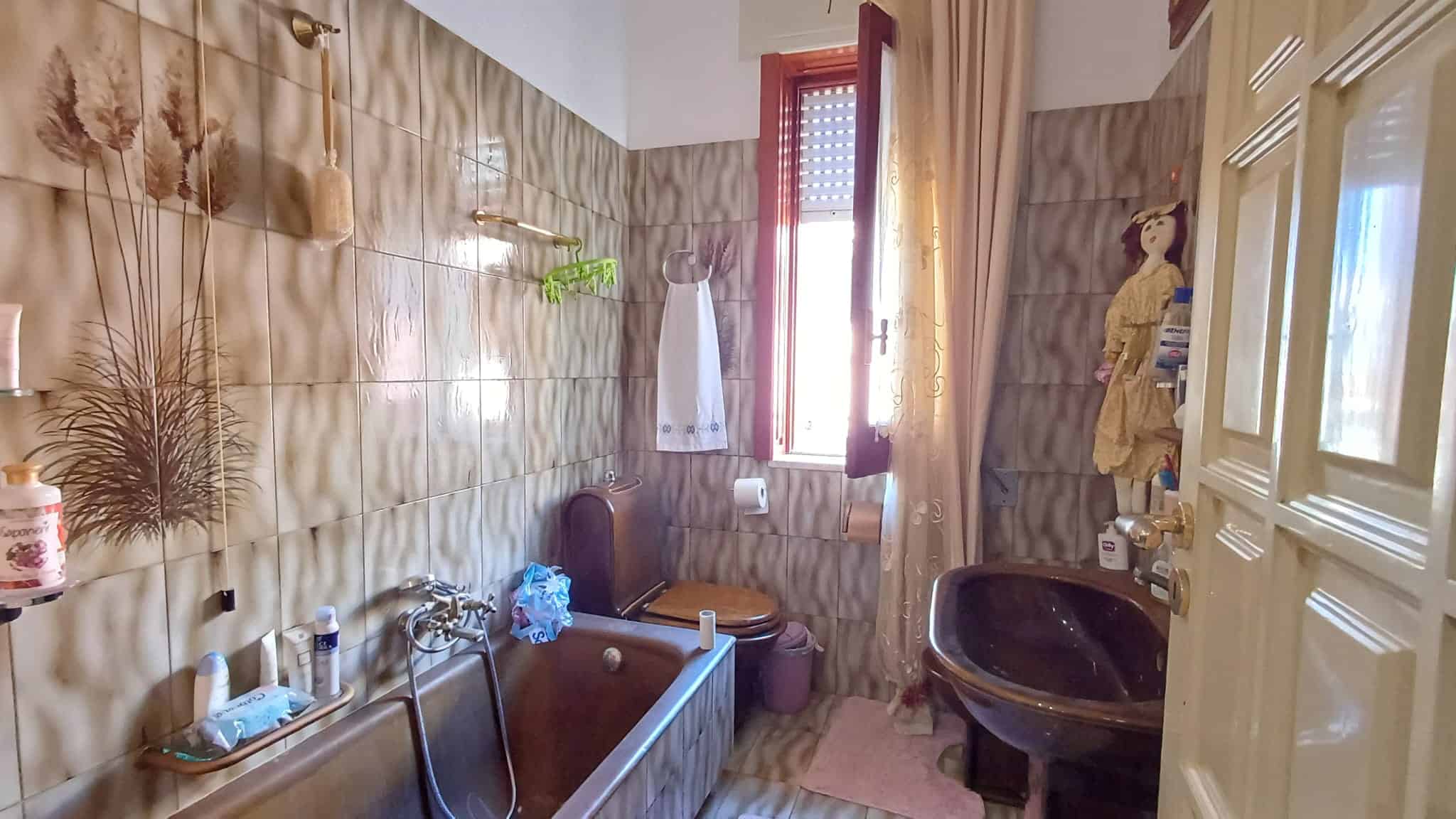 Ampia stanza da bagno con vasca, lavabo e finestra con tende, decorazioni floreali sulle pareti e mobili in stile classico, ideale per comfort e funzionalità.