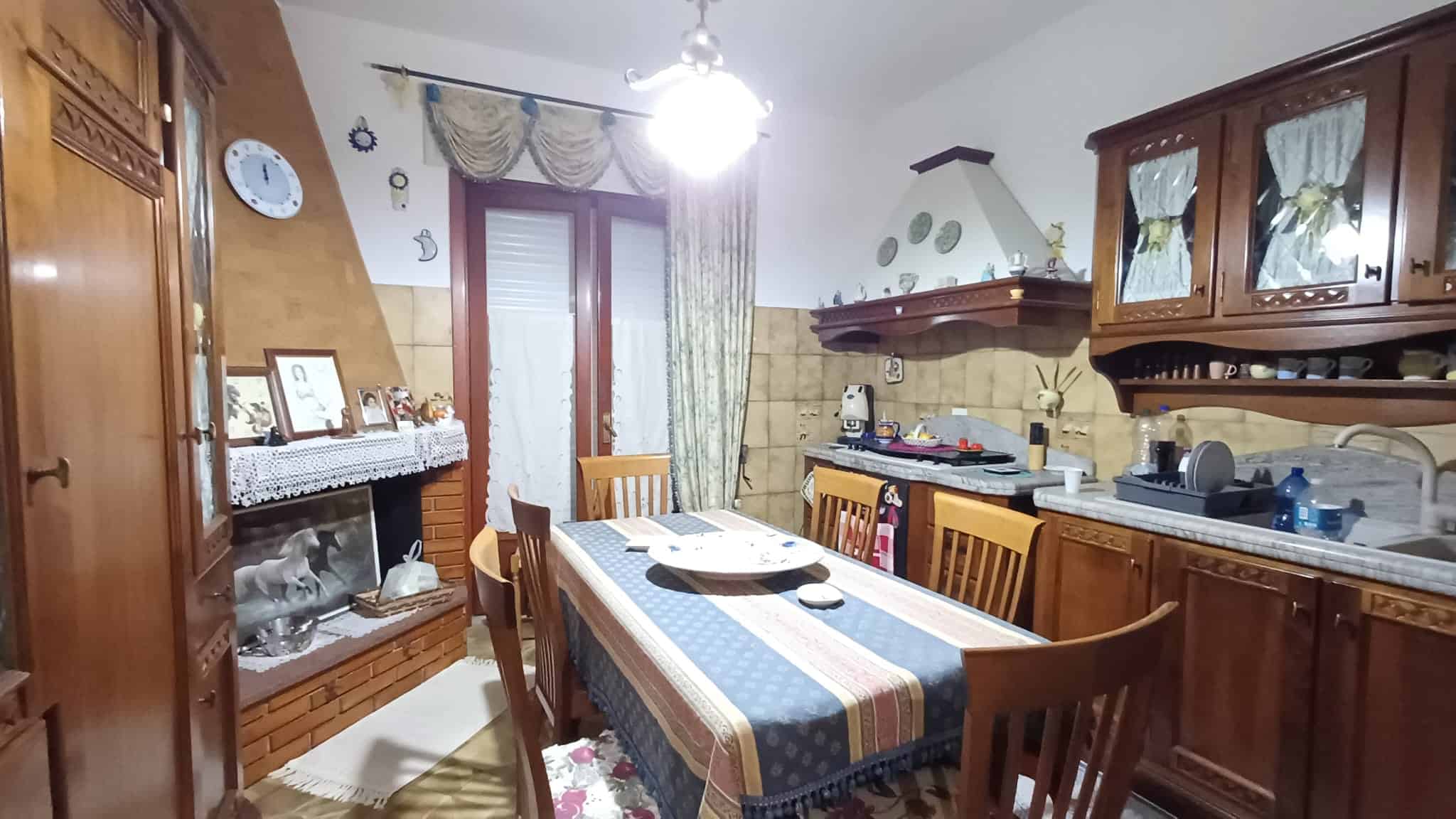 Una cucina tradizionale con mobili in legno, tavolo da pranzo con tovaglia blu e sedie in legno, ideale per famiglie, con elettrodomestici e decorazioni classiche.