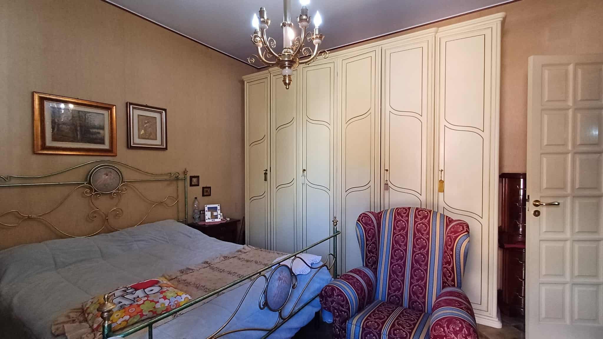 Armadio a muro bianco con ante a battente in una camera da letto elegante e luminosa, decorata con un letto matrimoniale, un comodino e un'illuminazione soffusa.