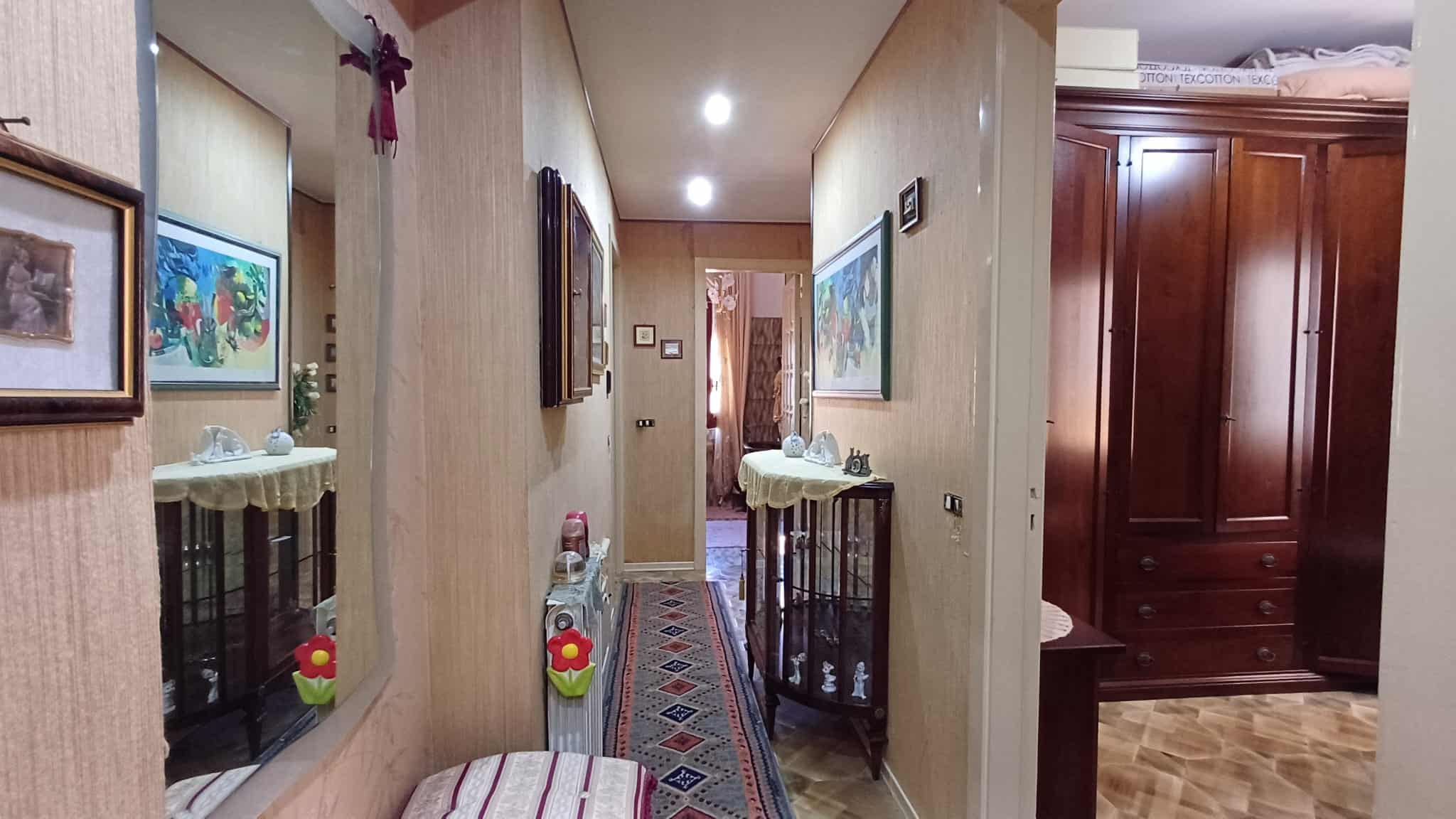 Elegante corridoio interno di una casa con pareti decorate, quadri alle pareti e un tappeto lungo al centro, illuminato da luci soffuse e decorazioni floreali, creando un ambiente accogliente e raffin.