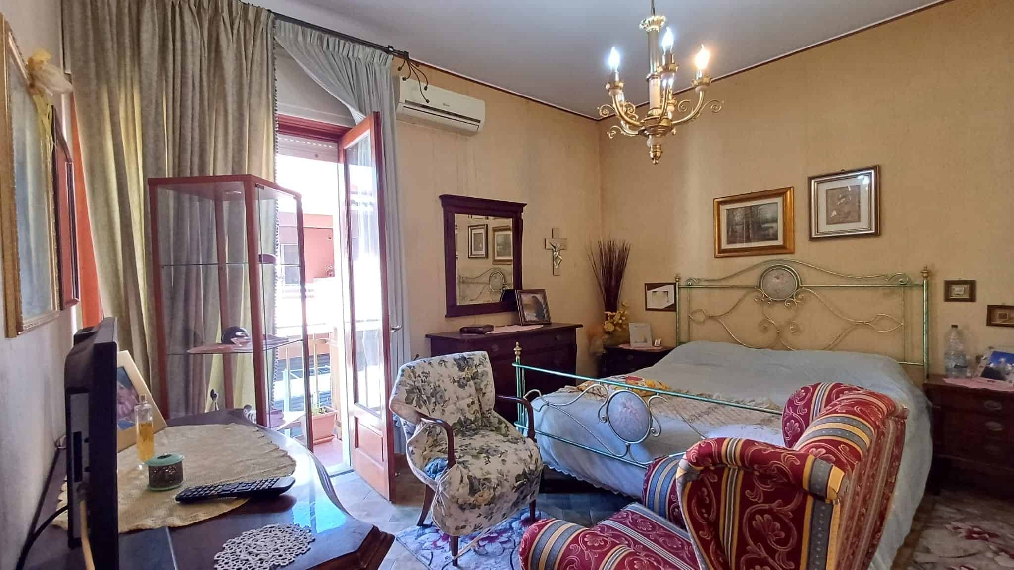 Ampia camera da letto con letto matrimoniale, sedie e mobili vintage, balcone con tende, decorazioni classiche, atmosfera accogliente e luminosa, ideale per soggiorni confortevoli.