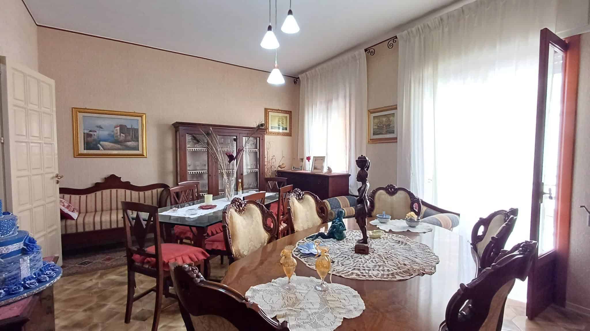 Interno di una sala da pranzo luminosa con tavolo in legno, sedie imbottite, tende leggere e decorazioni classiche, ambiente accogliente e ben illuminato.