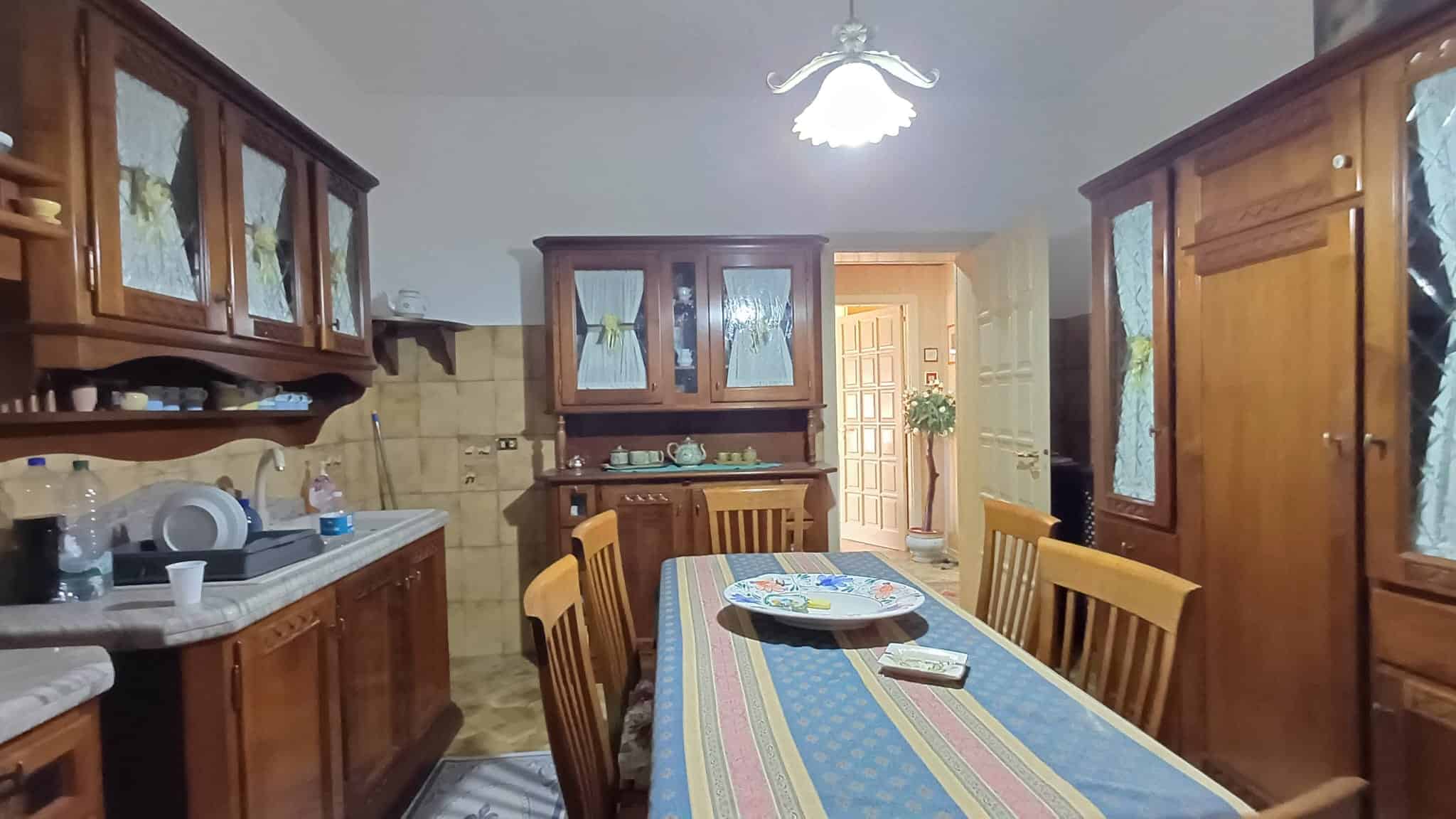 Pannello di cucina in legno con tavolo da pranzo, sedie e armadiature, ambiente domestico tradizionale con illuminazione centrale, dettagli di stoviglie e decorazioni in stile classico.