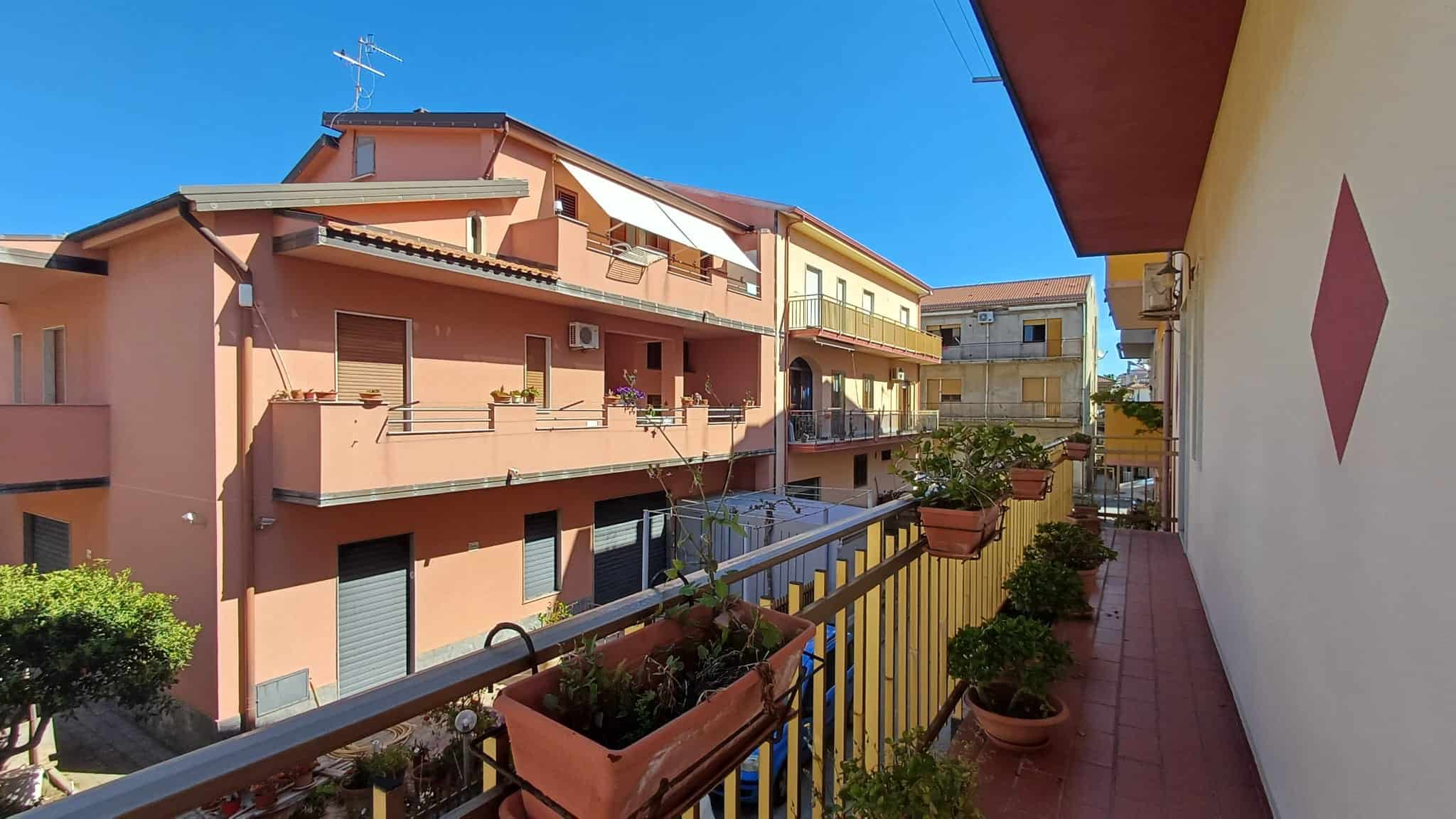 Balcone di un appartamento con piante in vasi, vista su edifici residenziali colorati e cielo azzurro, in un quartiere tranquillo.