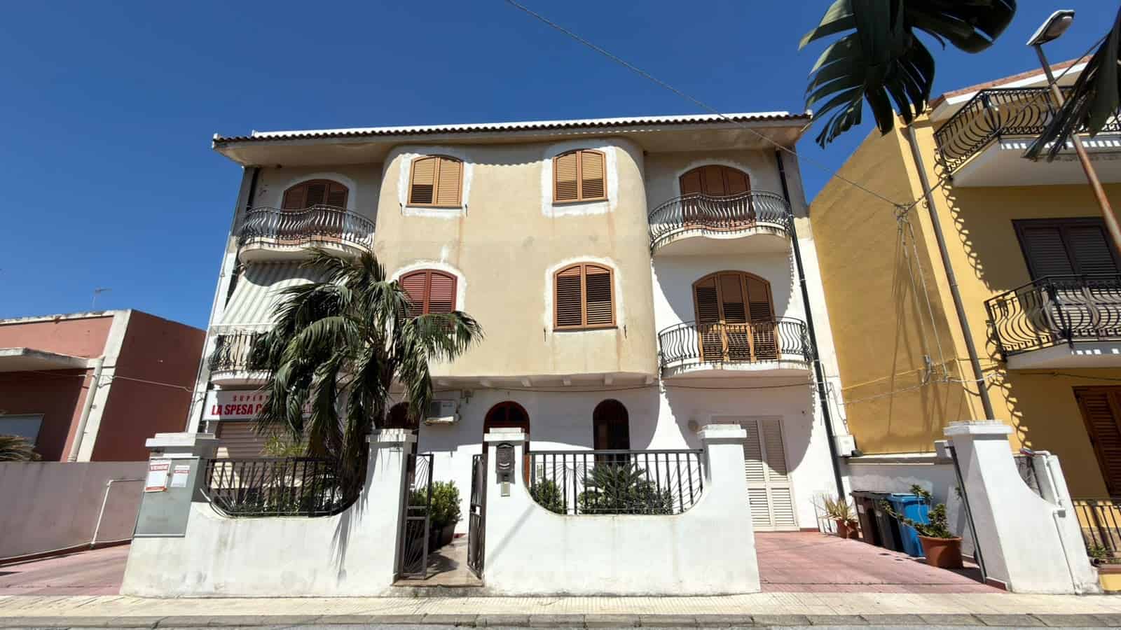 Elegante edificio residenziale con balconi in ferro battuto, pareti color crema e dettagli in legno, situato in una zona soleggiata con palme e piante tropicali, ideale per vivere in un ambiente tranq.
