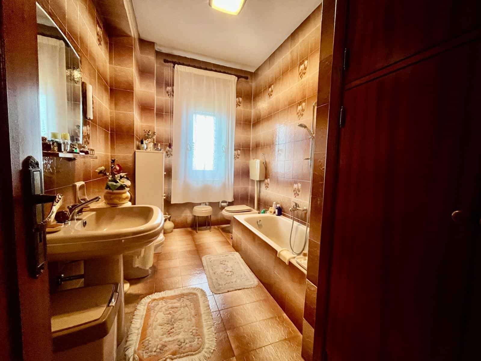 Lavabo e vasca da bagno in un bagno spazioso con pareti e pavimenti in piastrelle marroni, finestra con tende bianche, arredi eleganti e illuminazione calda, ideale per un'abitazione confortevole.