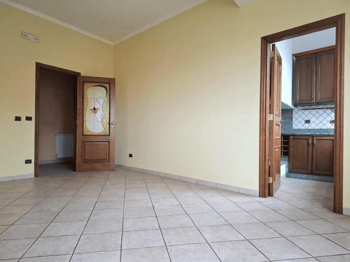 Ampio soggiorno vuoto con pareti gialle e pavimento in piastrelle, collegato a cucina con armadietti in legno e pareti rivestite di piastrelle blu, ideale per rinnovare e personalizzare gli spazi abit.