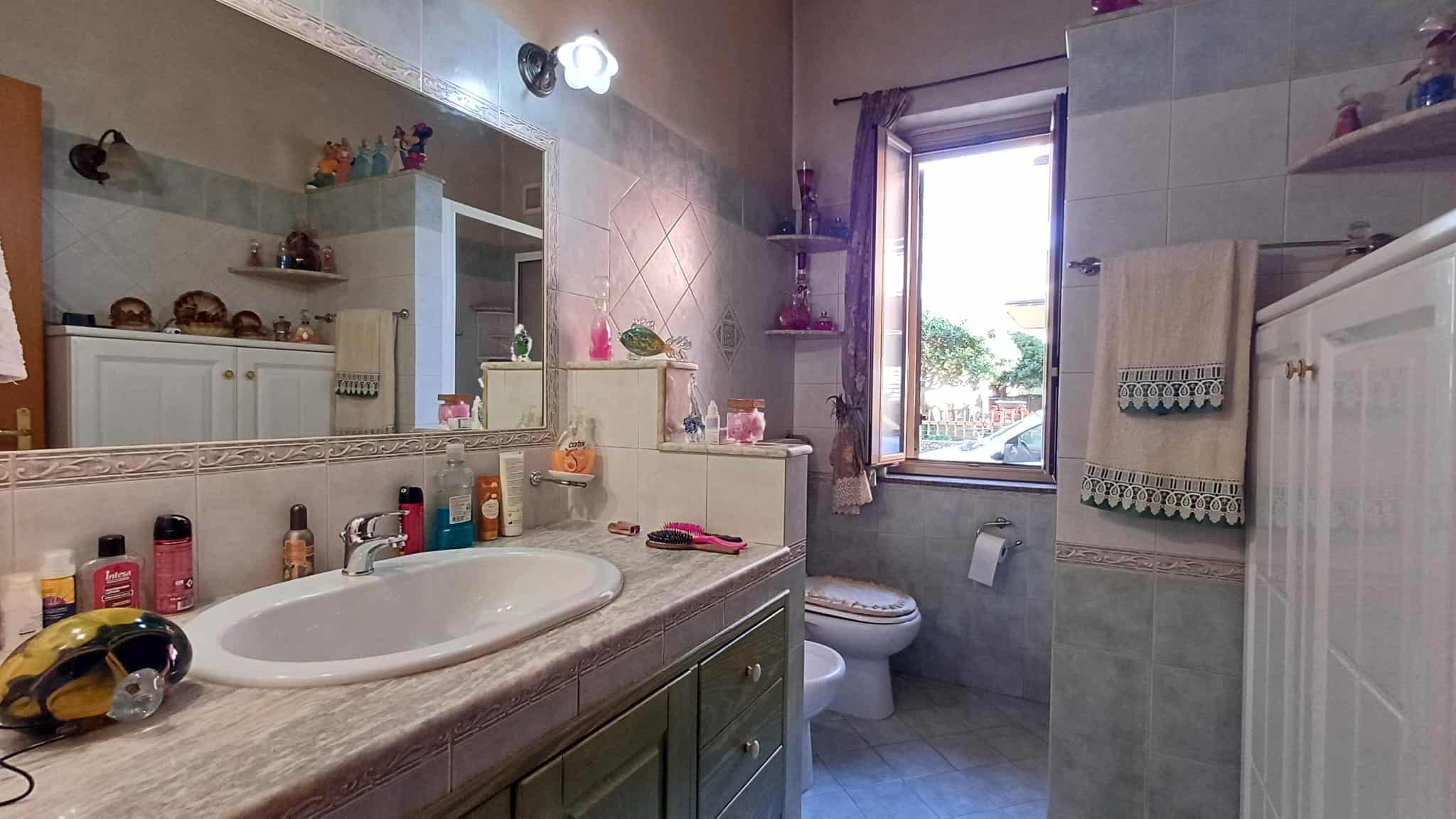 Lavabo e specchio in bagno con accessori e prodotti per la cura personale, finestra che permette l'ingresso di luce naturale, pareti rivestite in piastrelle chiare, ambiente accogliente e funzionale.