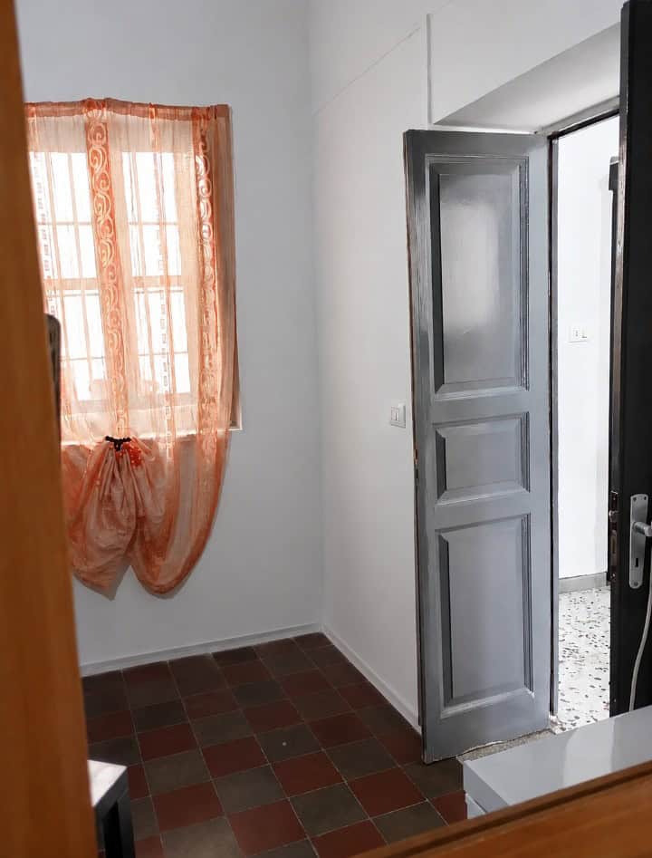 Interno di una stanza con pareti bianche, finestra con tende arancioni e porta aperta che conduce a un'altra stanza, pavimento in piastrelle rosse e grigie.
