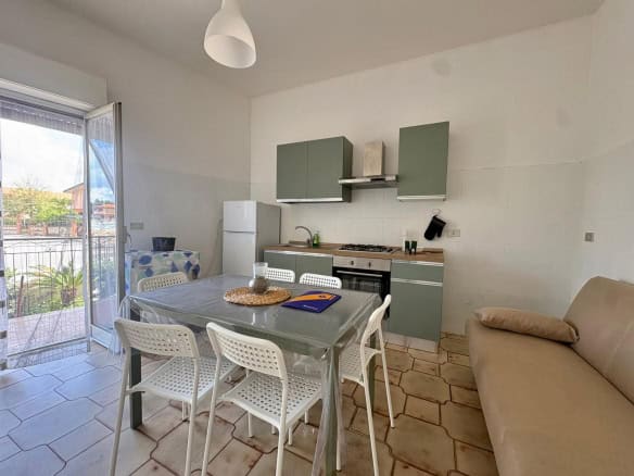 Interno di una casa moderna con cucina compatta e zona pranzo luminosa, con mobili minimalisti, tavolo con sedie bianche, e accesso a un balcone con vista all'aperto.