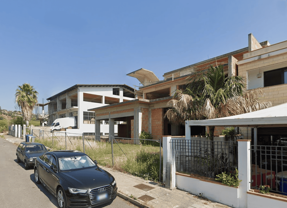 Villa moderna in costruzione con facciata in mattoni e ampi balconi, situata in zona residenziale con auto parcheggiate davanti.