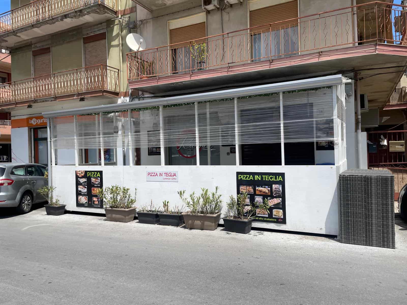 Pizzeria in teglia con area esterna coperta, menu visibile, piante decorative e sedie, situata in zona urbana.