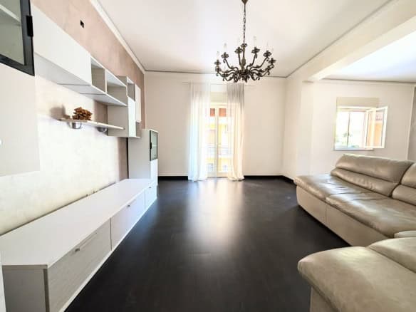 Interno di un soggiorno moderno con pareti chiare, divano beige, soffitto con lampadario e grandi finestre che lasciano entrare molta luce naturale.