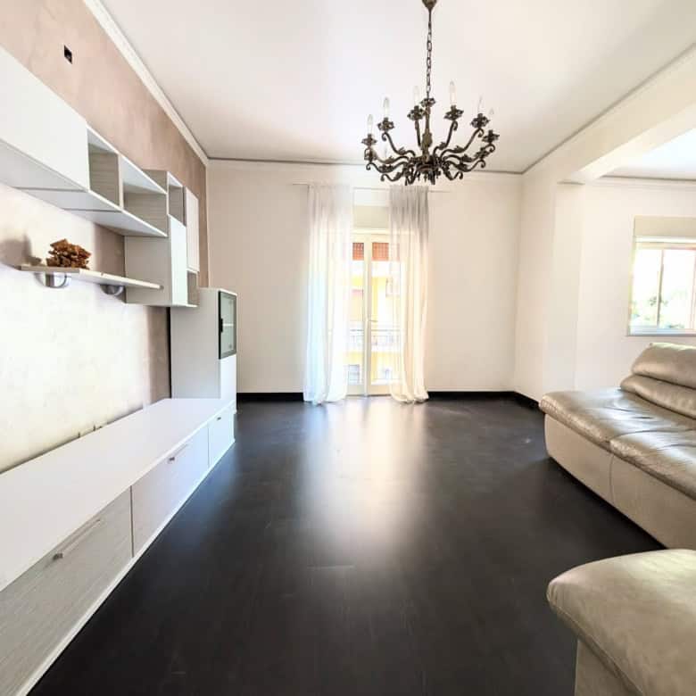 Interno di un soggiorno moderno con pareti chiare, divano beige, soffitto con lampadario e grandi finestre che lasciano entrare molta luce naturale.