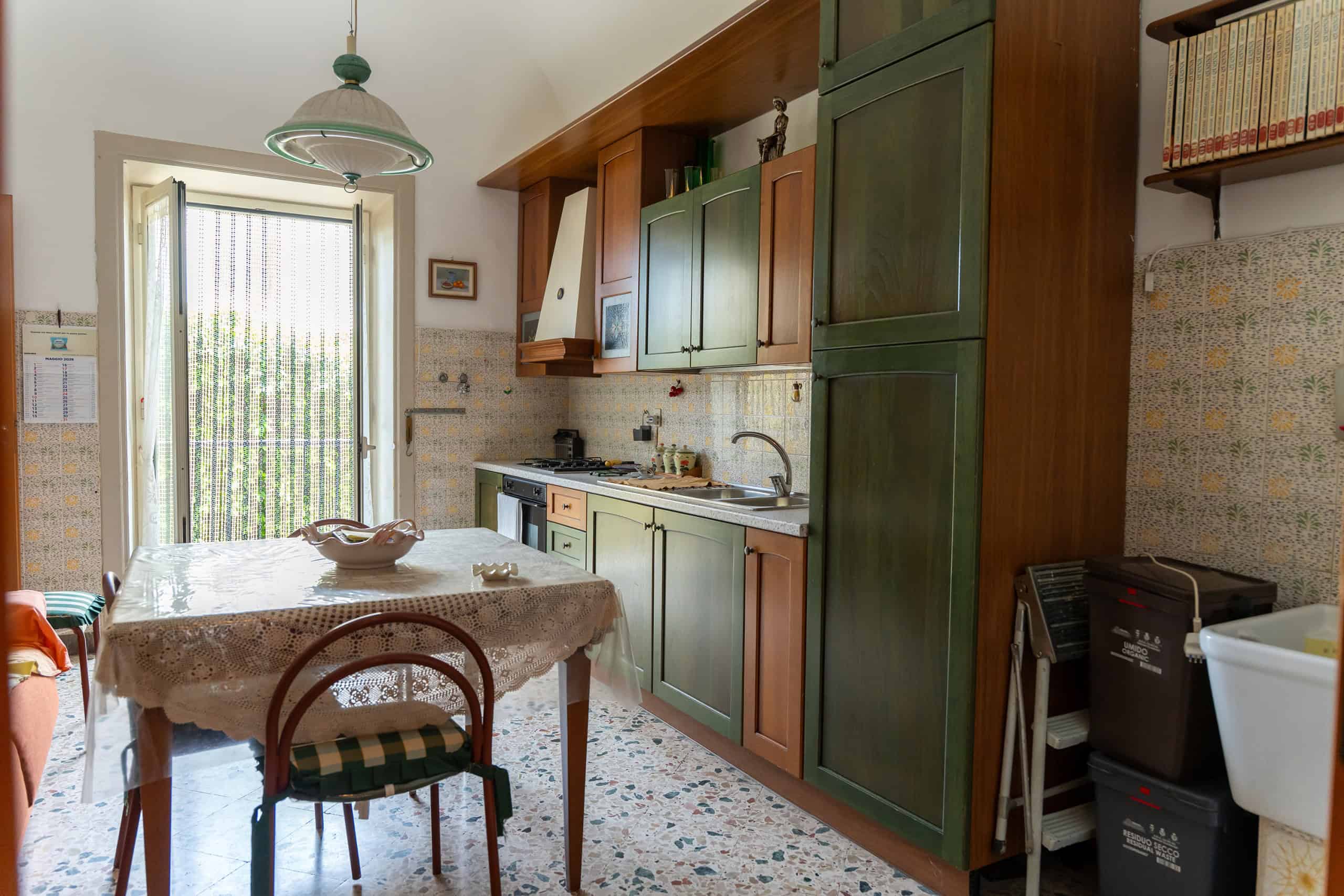 Una cucina abitabile con mobili in legno verde e beige, tavolo da pranzo con tovaglia ricamata, e una porta finestra che permette l'ingresso di luce naturale.