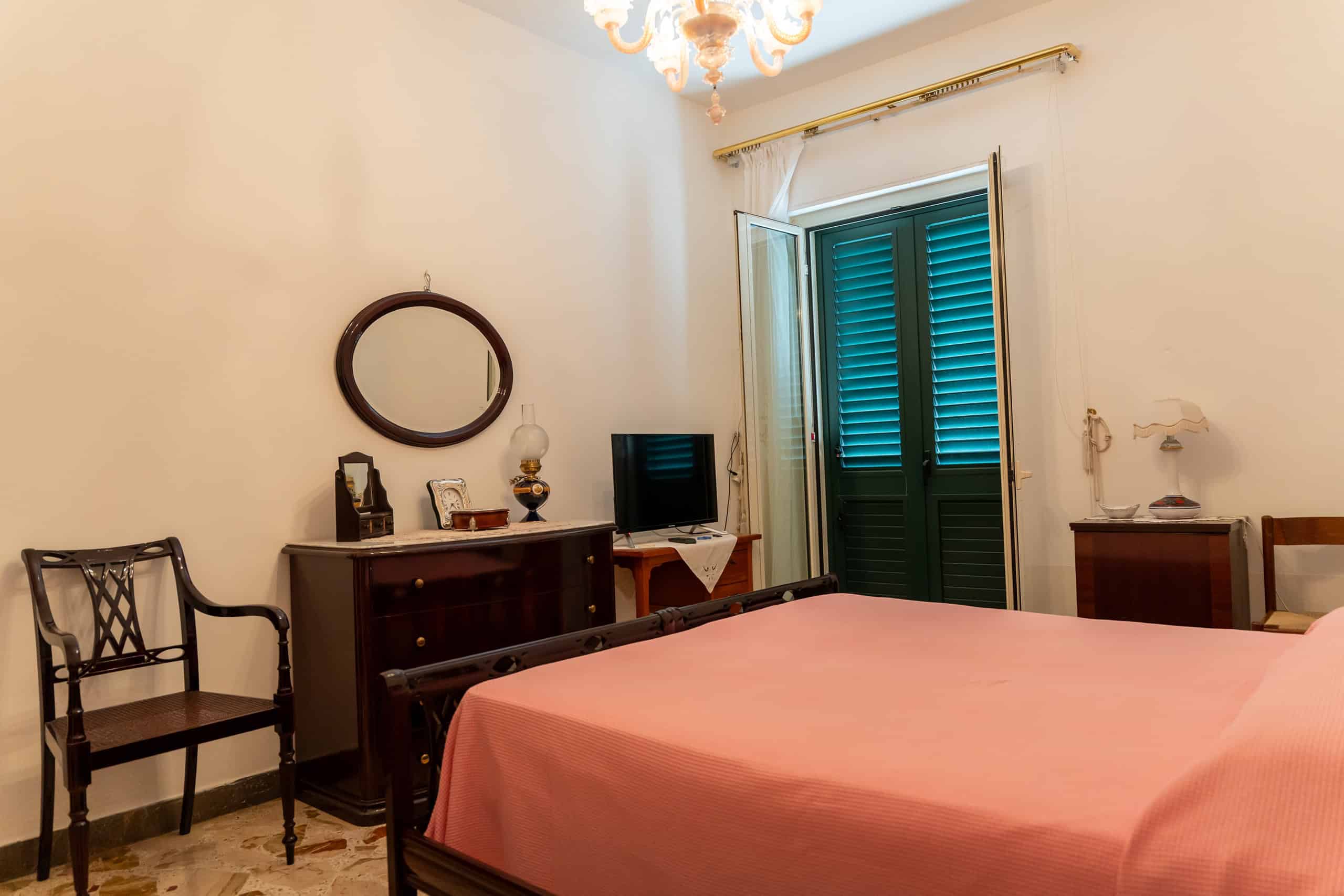 Un'immagine di una camera da letto con pareti bianche, un letto con copertura rosa, un armadio, una sedia in legno e un televisore, illuminata da una lampada e una finestra con persiane verdi.