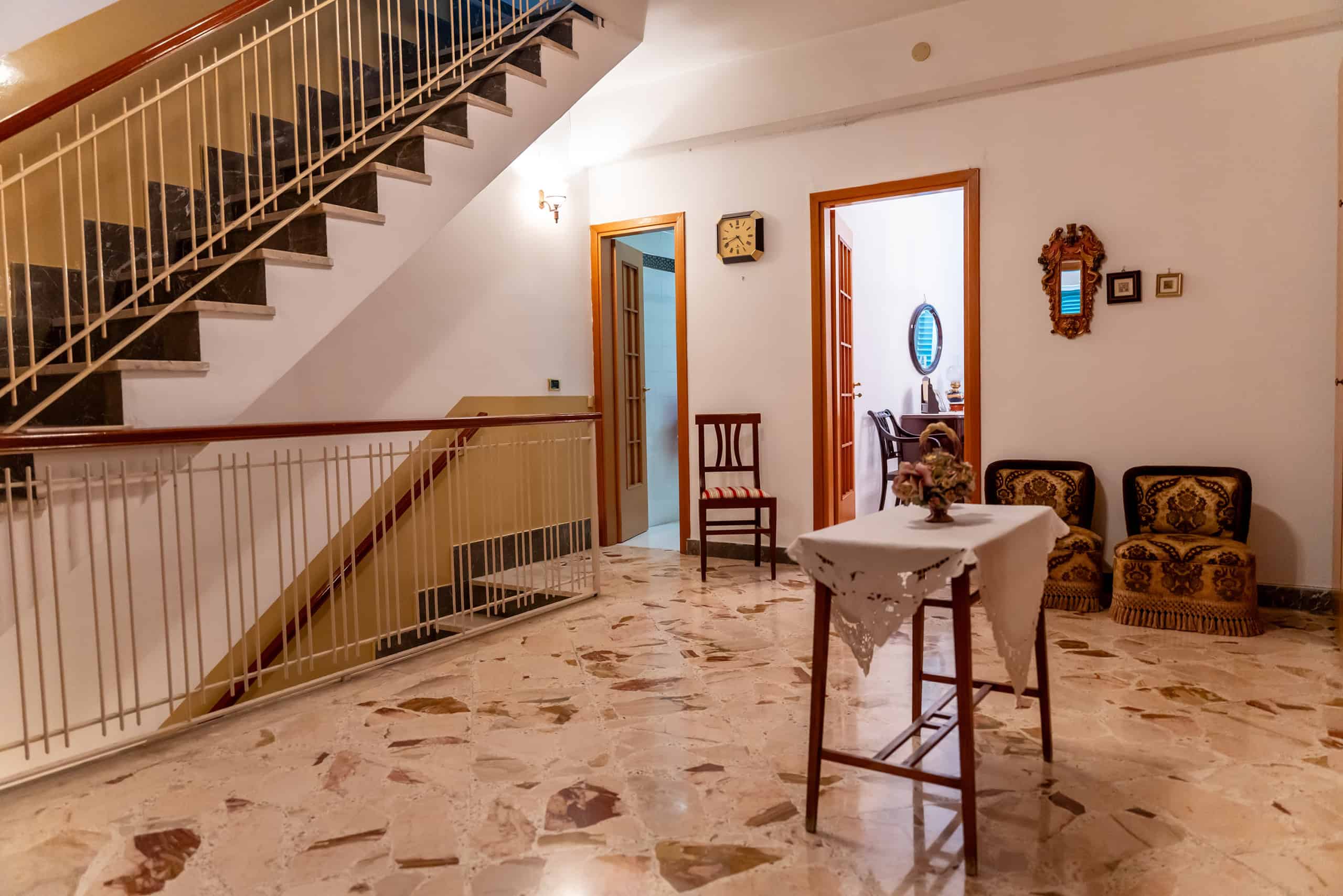 Interno di una casa con scala, tavolo decorato e sedie, arredamento classico, pavimento in marmo, pareti bianche, stile accogliente e tradizionale.