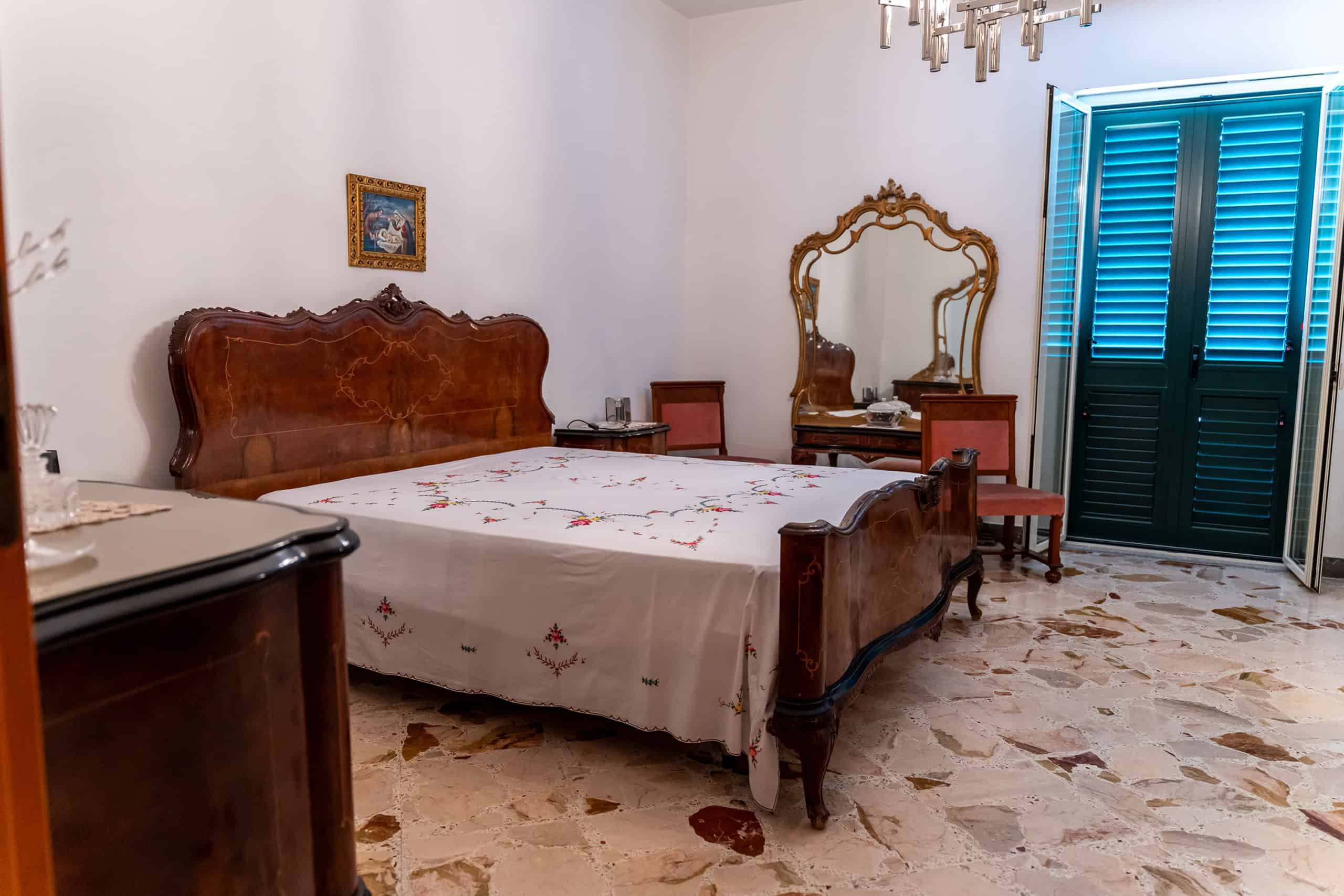 Una camera da letto con arredi classici, letto in legno con testiera intagliata, specchio antico e finestra con persiane blu, pavimento in marmo chiaro.