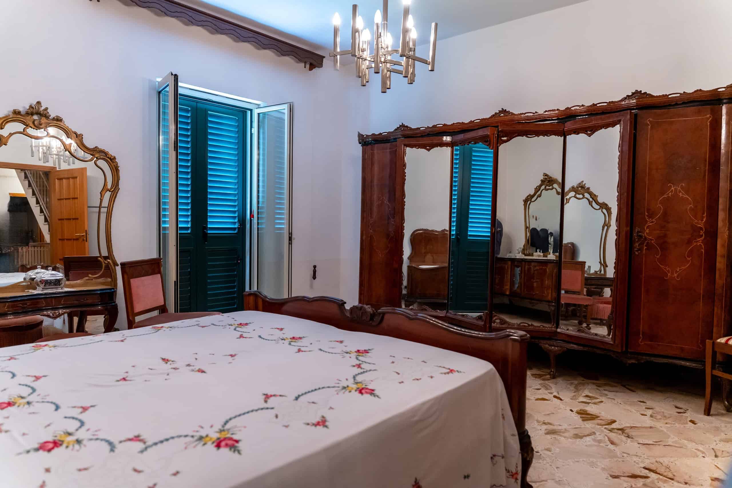 Anticamera da letto con armadio in legno e specchi, finestra con persiane blu, arredamento classico, lampadario a soffitto, pavimento in marmo, stile elegante e tradizionale.