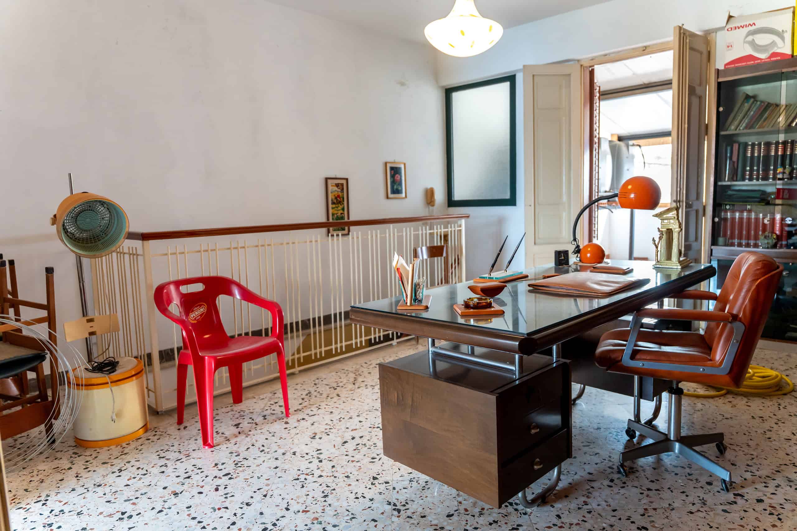 Un ufficio domestico con scrivania, sedie e libreria, illuminato dalla luce naturale, con pareti bianche e pavimento in marmo, ideale per lavoro e studio.