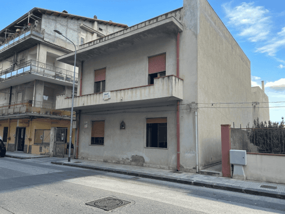 Ampio edificio residenziale in fase di ristrutturazione con facciata in cemento e finestre con tapparelle rosse, situato in zona urbana tranquilla con strada asfaltata e marciapiede.