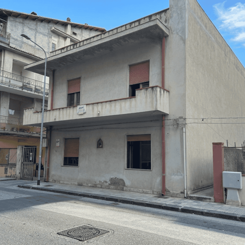 Ampio edificio residenziale in fase di ristrutturazione con facciata in cemento e finestre con tapparelle rosse, situato in zona urbana tranquilla con strada asfaltata e marciapiede.