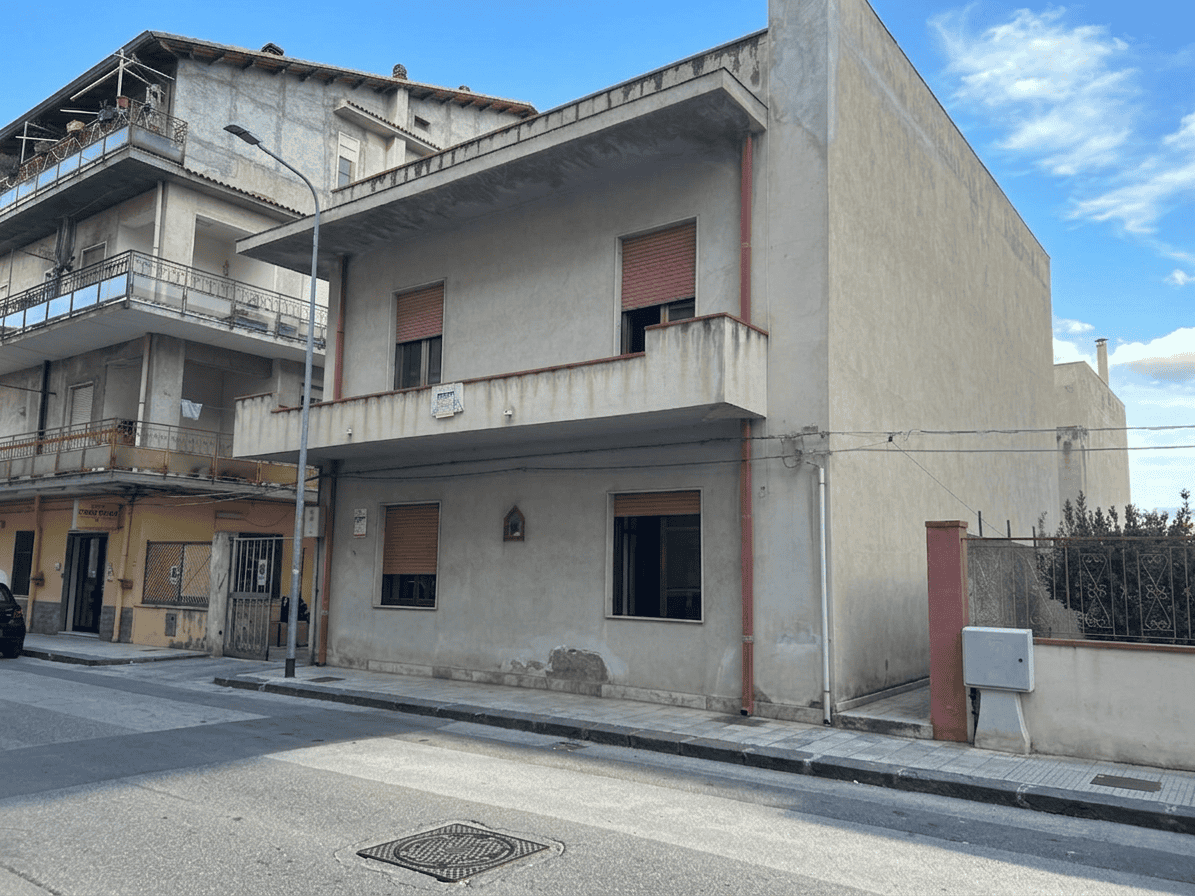 Ampio edificio residenziale in fase di ristrutturazione con facciata in cemento e finestre con tapparelle rosse, situato in zona urbana tranquilla con strada asfaltata e marciapiede.