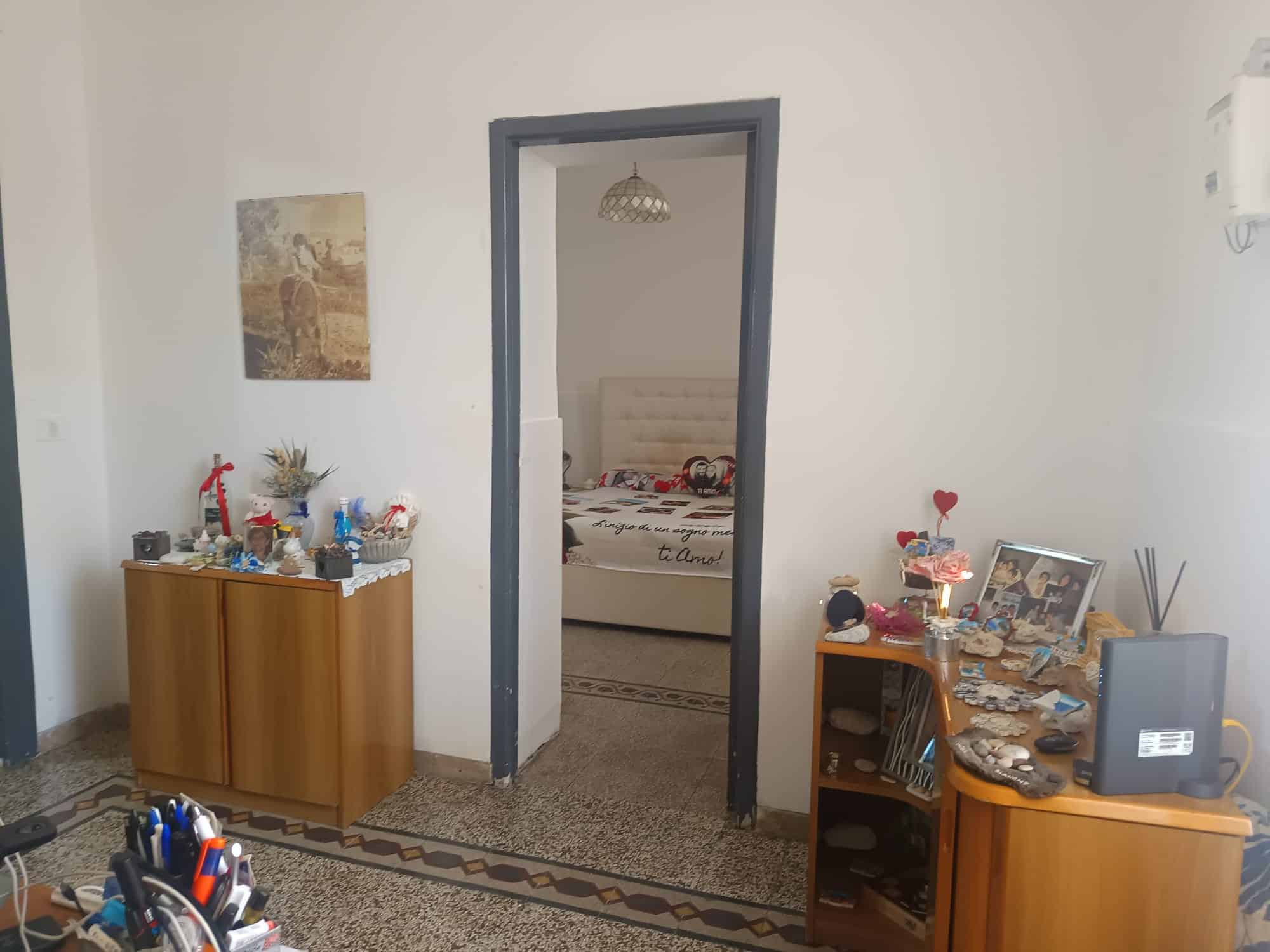 Interno di una casa con mobili in legno, decorazioni e un ingresso che conduce a una camera da letto con testiera imbottita e decorazioni romantiche.