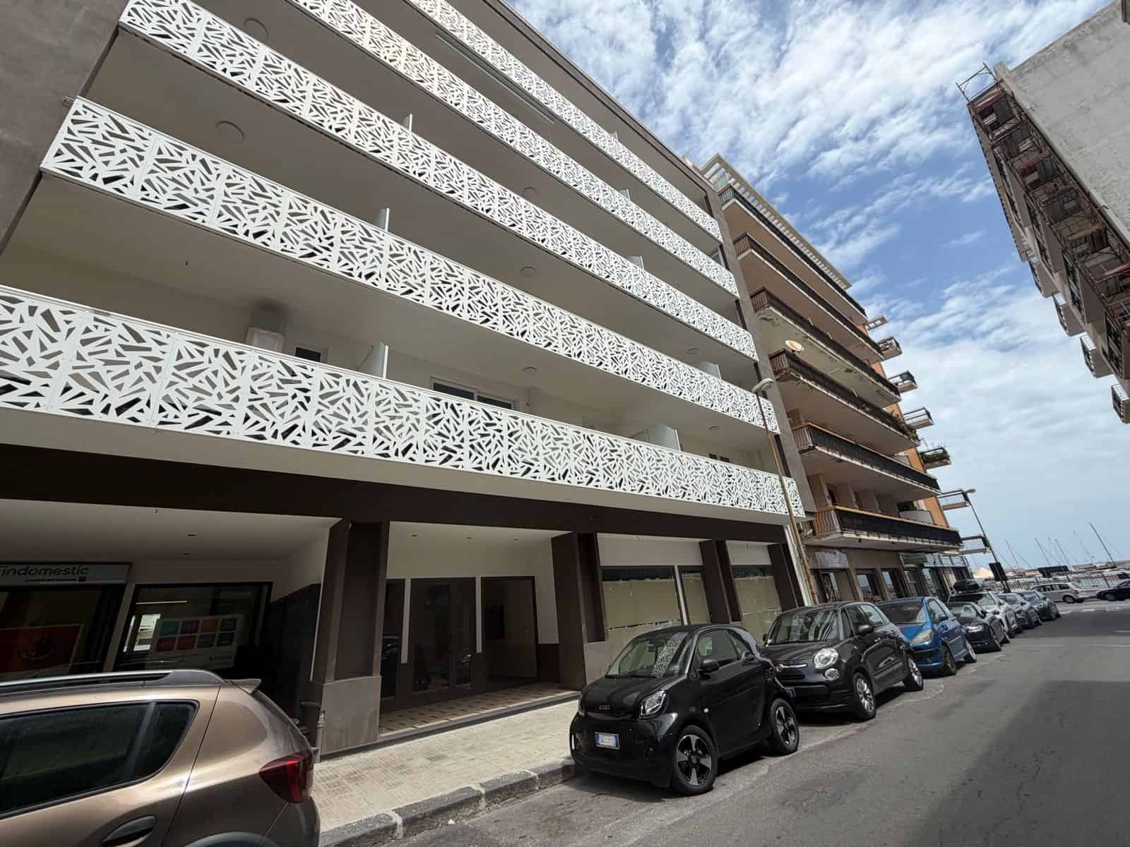 Balcone di un edificio moderno con dettagli decorativi in stile geometrico, parcheggio con auto lungo la strada, cielo nuvoloso, zona urbana in fase di sviluppo.