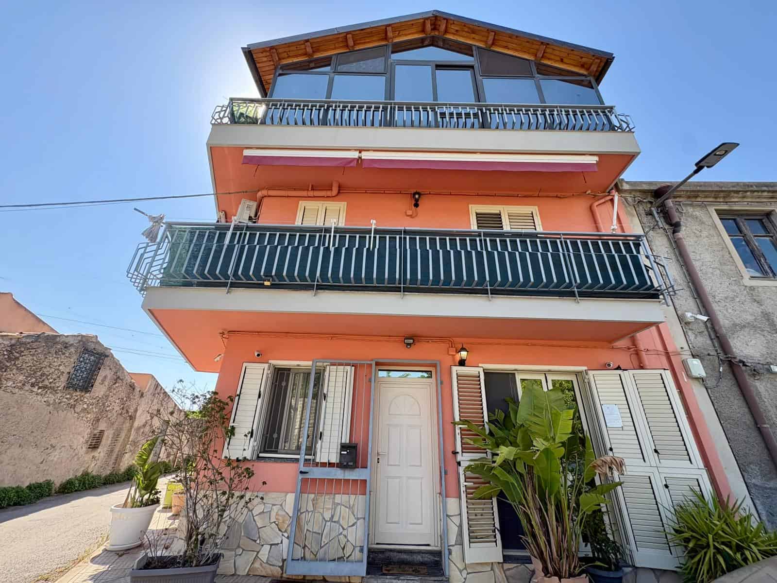 Ampio edificio residenziale a tre piani con balconi e grandi finestre, situato in zona urbana soleggiata, con piante ornamentali e accesso pedonale. Ideale per investimento immobiliare o residenza pri.
