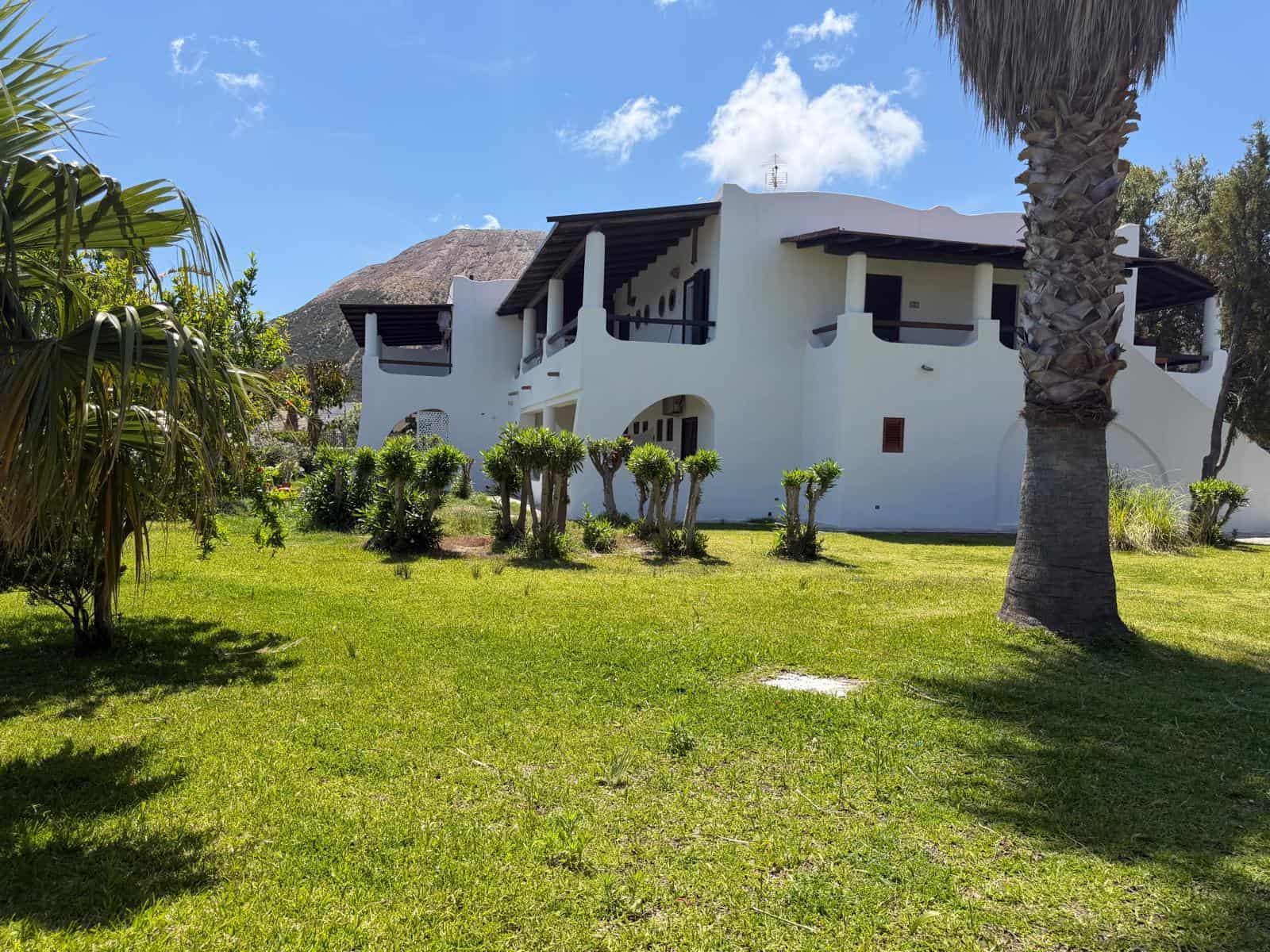 Ampia casa moderna bianca con terrazza e balconi, immersa in un giardino verde con palme e piante ornamentali, sotto un cielo azzurro con nuvole, in un ambiente tropicale.