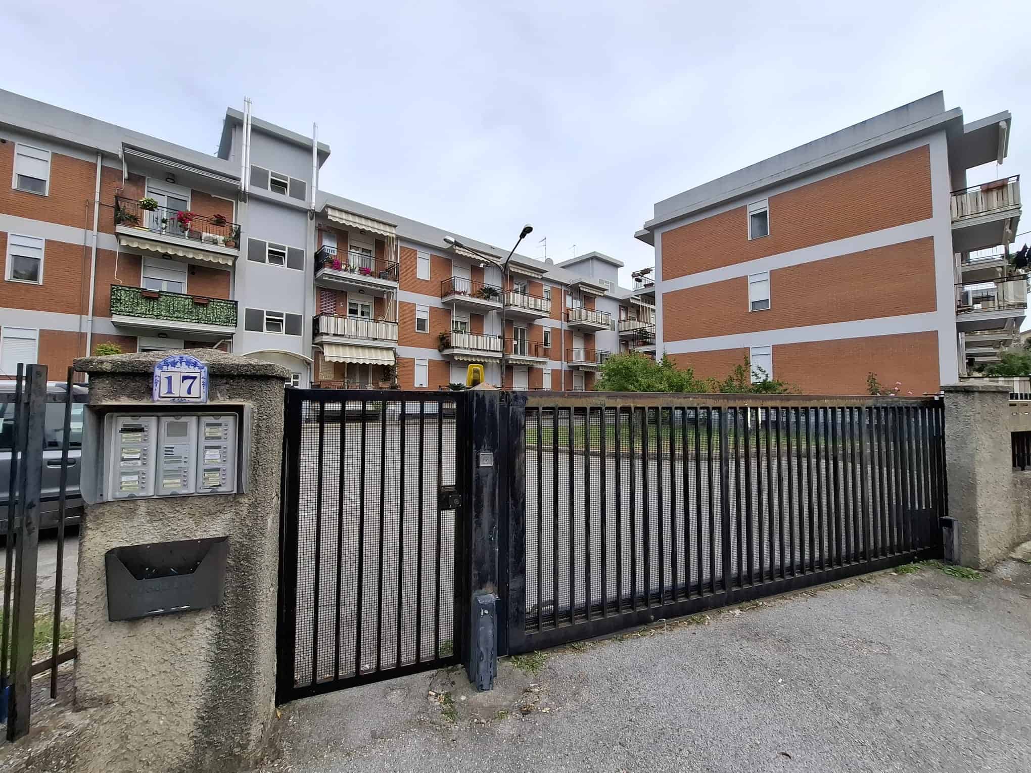Cancello in ferro nero con pilastri in cemento, ingresso di un complesso residenziale con appartamenti moderni e balconi fioriti, in un quartiere urbano di città italiane.