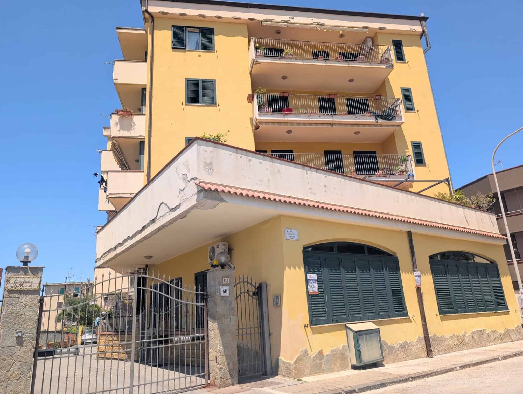 Ampio edificio residenziale di quattro piani con balconi e finestre, facciata gialla e ingresso con cancello in metallo, situato in zona urbana soleggiata.